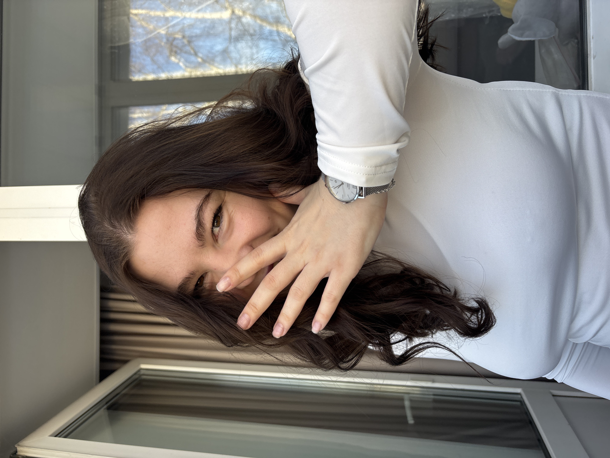 The image depicts a close-up, chest-up view of a young adult female situated indoors in the city of Perm, Russia. She has long, dark brown wavy hair that frames her face and dark brown eyes, which are visibly smiling. Her right hand, with natural and unpolished nails, is raised and covers her mouth and nose. She wears a form-fitting white long-sleeved top and a silver-toned wristwatch with a mesh strap and a white dial on her left wrist.
The indoor environment includes a partially open, white-framed glass door or large window to the left, with closed brown vertical blinds or curtains visible behind it. To the right, another white-framed window provides a view of the outdoors, showcasing a clear blue sky and numerous bare tree branches, consistent with birch trees in a late winter or early spring season. A blurred white and yellow object is partially visible through the lower right pane of this window.