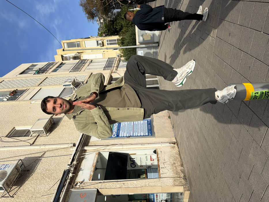 A male individual stands centrally on a paved Tel-Aviv sidewalk, balancing his right foot on a gray cylindrical bollard marked with a yellow reflective stripe and partially visible Hebrew text, including "גישה שווה לכל" (Equal access for all). He faces forward, wearing a light olive-green long-sleeved shirt over an olive-green t-shirt, dark gray trousers, and white athletic shoes with green accents. His left leg is bent with the foot raised, and his hands are pressed together in a prayer-like pose at chest level. He looks directly at the camera with a slight smile.
Behind the subject, a multi-story building with a textured beige facade displays several window-mounted air conditioning units. Its ground floor features a commercial establishment with a window displaying "GMT" and "MoneyGram" logos, and an adjacent blue sign with "CHANGE" in English, accompanied by extensive Hebrew text detailing services. To the right, another male individual with dark skin is partially visible from the back, wearing a dark denim jacket and black pants, appearing to interact with a mobile phone. Further in the background, additional multi-story buildings and trees are visible under a clear blue sky.