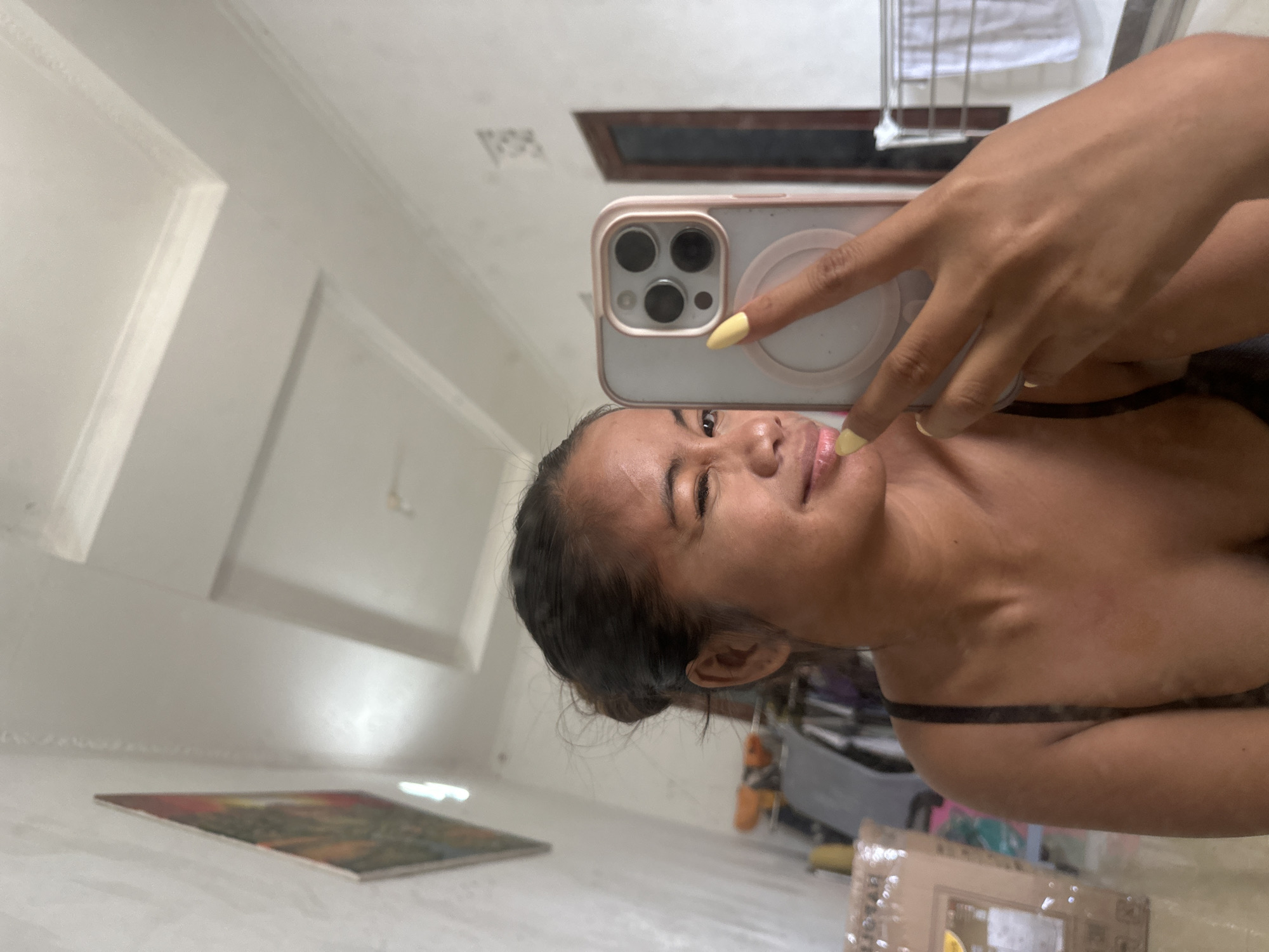 A young woman with dark hair tied back is taking a selfie in a mirror. She is winking and smiling, wearing a dark camisole. Her right hand is visible, holding a phone with a light pink case. The background shows a room with a white ceiling with decorative molding, a framed picture on the wall, clothing racks, and a drying rack with towels. The overall impression is a casual snapshot taken in a domestic setting, likely a bedroom or bathroom in Sukawati, Indonesia.
