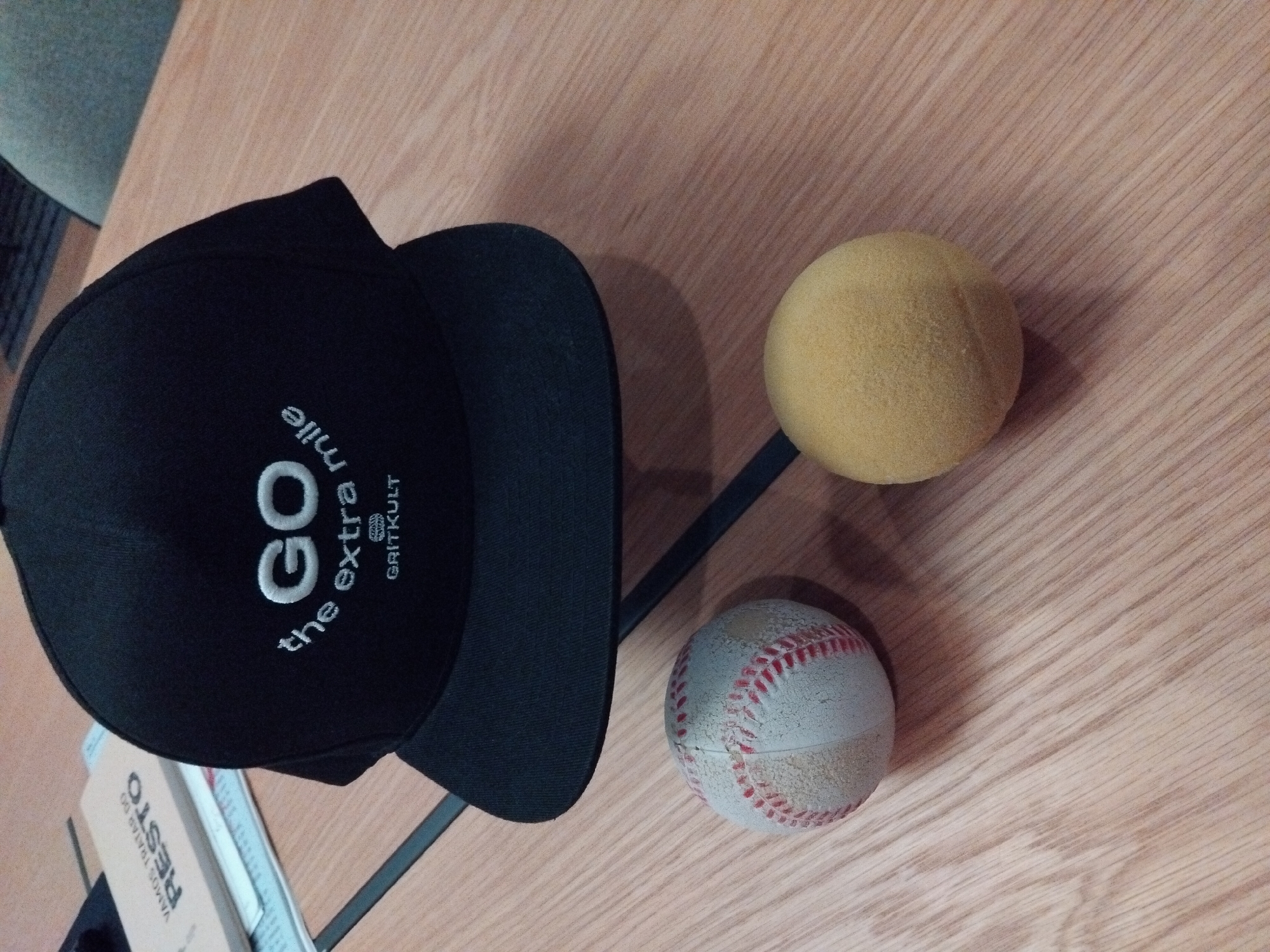 A black baseball cap with white embroidery sits on a wooden table, its brim pointing towards the viewer. The embroidery on the cap reads "GO the extra mile" in a curved fashion, with "GRITKULT" below it. To the left of the cap, a worn baseball with red stitching rests on the table. To the right of the cap, a soft, yellow ball is also on the table. In the background, a portion of a book with text that appears to say "VAMOS TRATAR DO RESTO" is visible. The setting is an indoor environment with warm lighting. This image was taken in Vila Nova de Famalicão, Portugal.