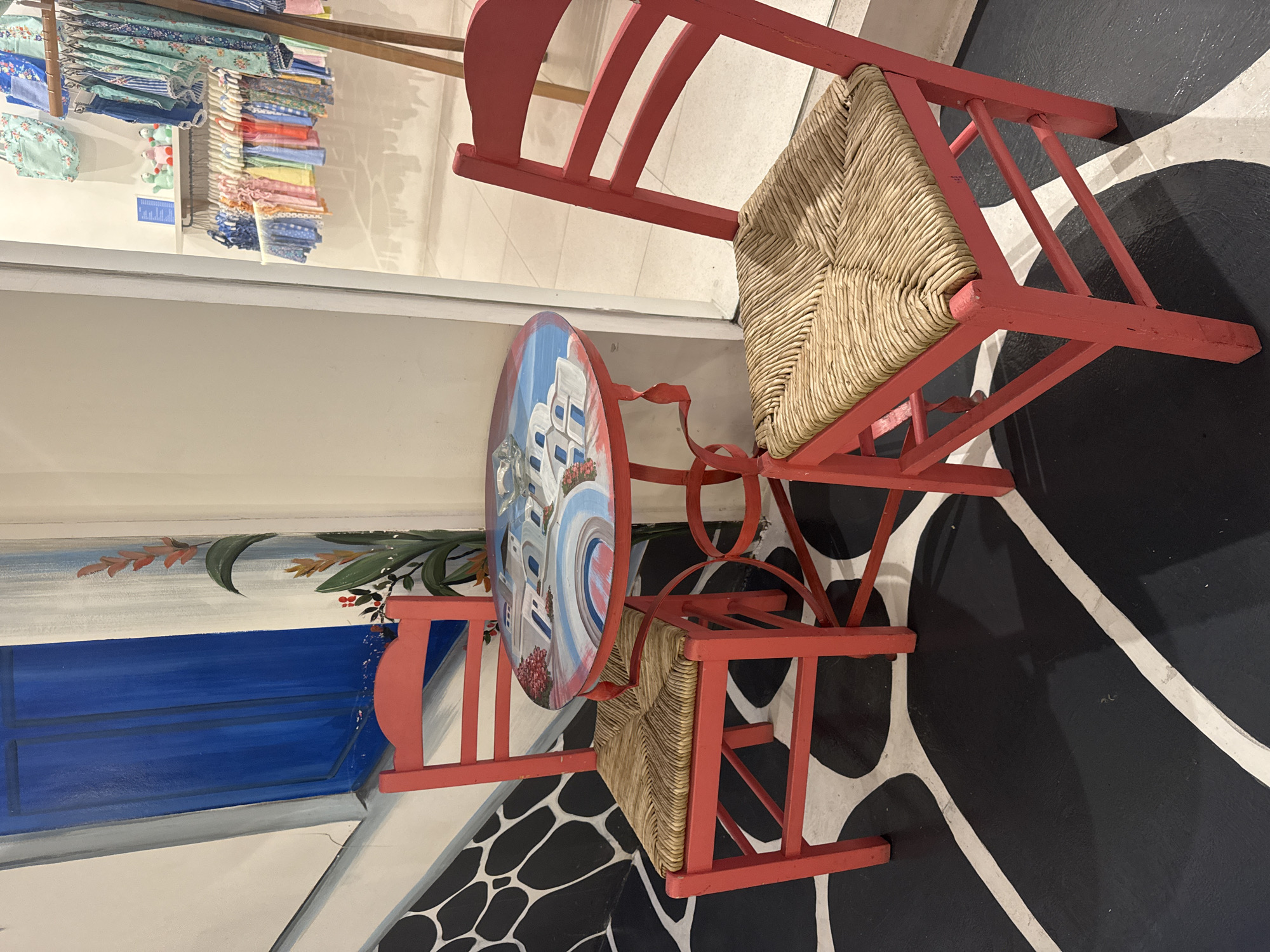 A small table and two chairs, all painted red, are placed on a black and white patterned floor, likely in a shop in Denpasar, Indonesia. The chairs have woven straw seats. The table has a round top painted with a scene of a blue and white building, possibly representing Santorini, Greece. To the left, a mural shows a blue window, and to the upper left, clothing is displayed on a rack.