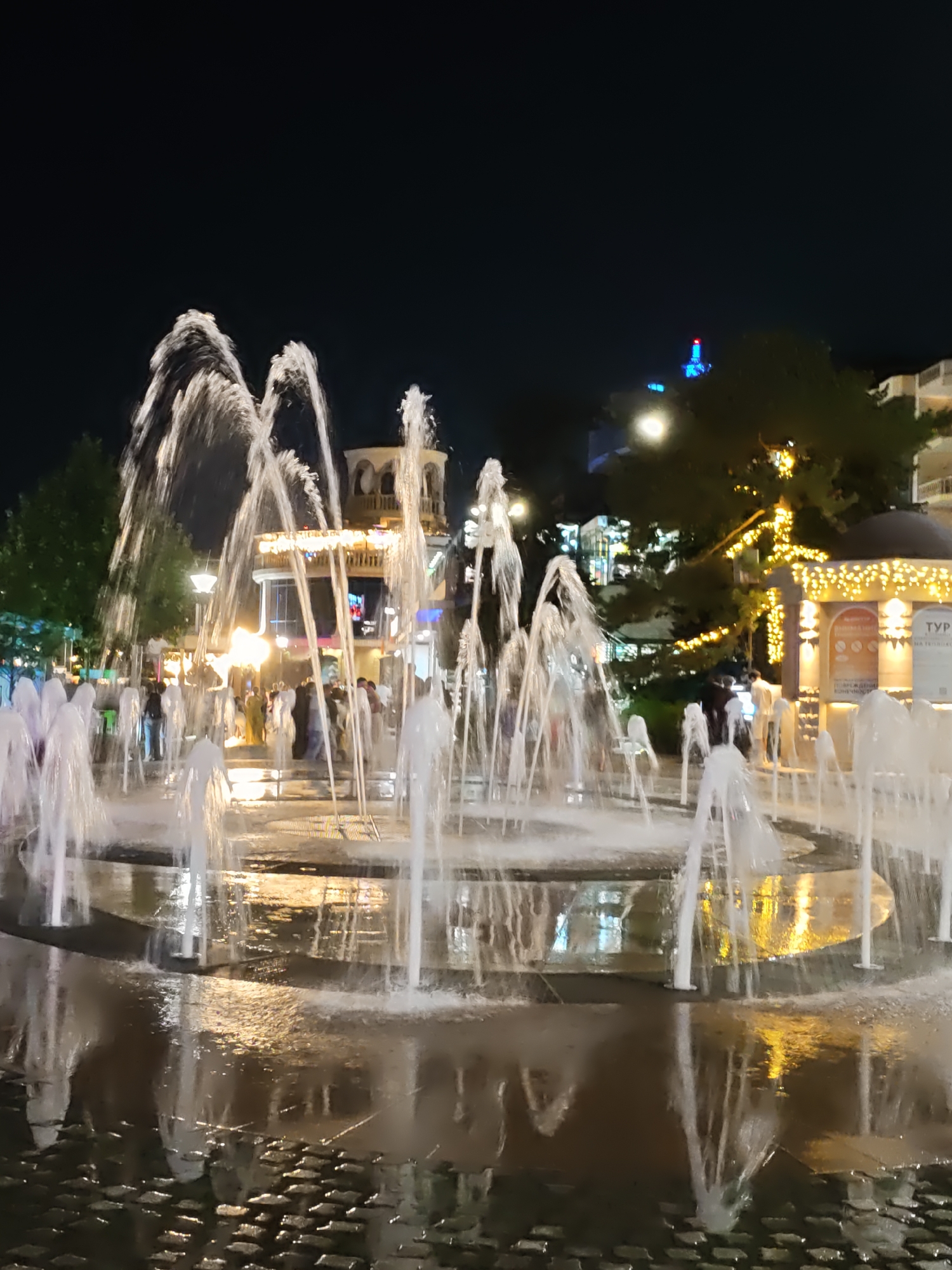 A fountain plaza is depicted at night in Rostov-on-Don, Russia, with water jets illuminated and reflecting on the wet, paved ground. In the background, a building with illuminated arches and decorative lights is visible, along with trees adorned with string lights. Several people are present, some interacting in groups, and others walking through the plaza. The overall atmosphere is festive, with the night sky providing a dark backdrop to the bright lights and water features.