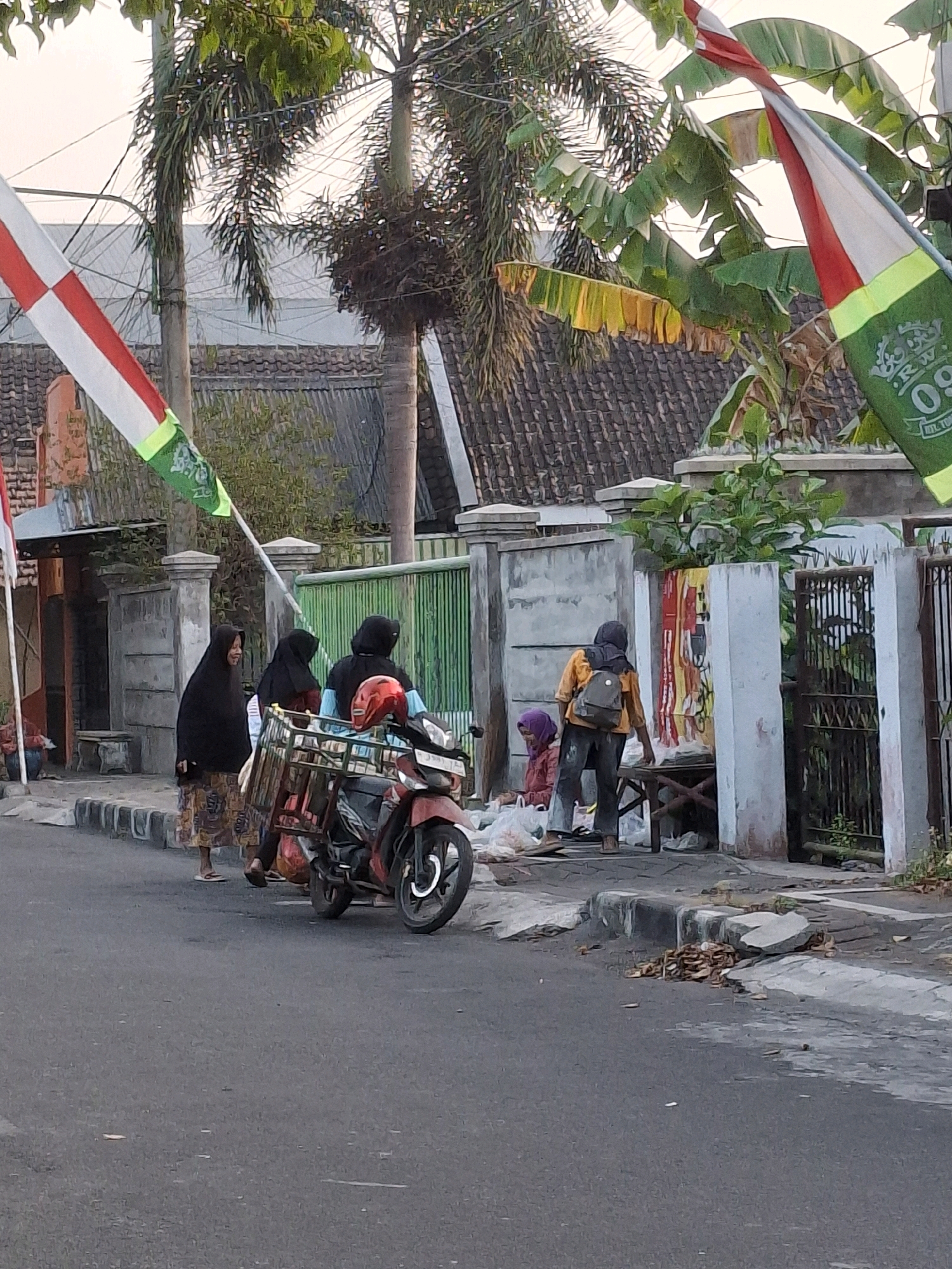 In Lumajang, Indonesia, on a paved road, a group of people are gathered near a sidewalk. A red motorcycle with a metal basket attached to the back is parked. Three women in hijabs are near the motorcycle. One woman in a patterned skirt and black hijab is standing. Two other women, one with a blue shirt visible, are standing behind the motorcycle. A red helmet rests on the motorcycle. Further down the sidewalk, another person wearing an orange shirt and backpack is interacting with a person seated in a pink outfit, surrounded by plastic bags. The scene is framed by trees, houses with tiled roofs, and flags on poles. A metal gate is visible in the background.