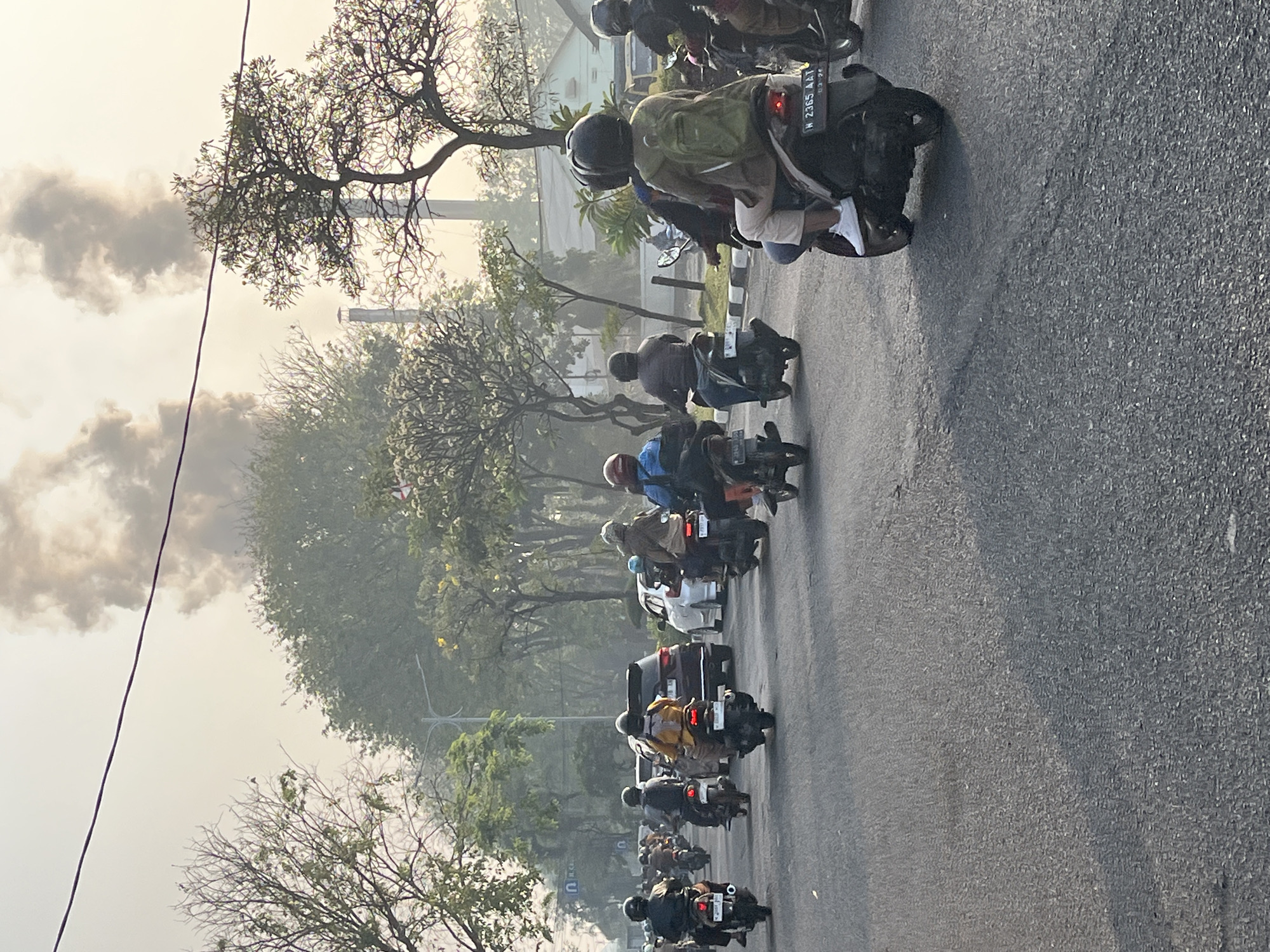 The image shows a street scene in Candi, Indonesia, characterized by heavy traffic and industrial pollution. A multitude of motorcycles, along with a few cars, are navigating a paved road. Many of the motorcyclists are wearing helmets and some are carrying backpacks. In the background, large plumes of dark smoke are rising into the hazy sky, originating from industrial smokestacks. Trees line the sides of the road, with some foliage appearing lighter due to the atmospheric conditions. The overall impression is one of urban life impacted by industrial emissions.
