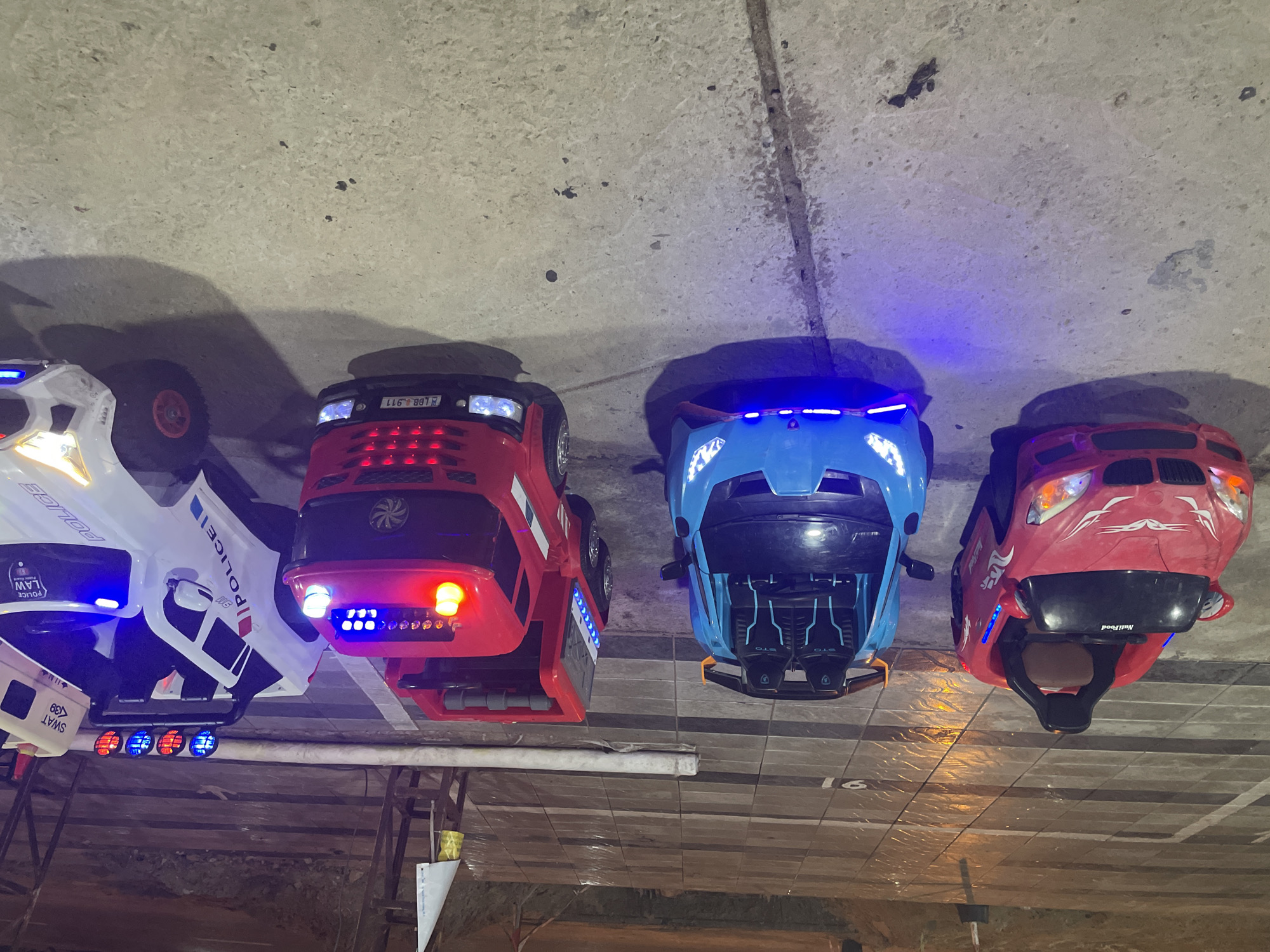 The image shows four children's ride-on toy cars parked indoors, presumably in a commercial or entertainment area. The cars are lined up and feature a white police car, a red fire truck, a blue sports car, and another red car. All the cars have illuminated lights. The location is possibly in Mũi Né, Vietnam, based on external context. The vehicles are parked on a tiled surface, with visible concrete or ceiling structure above. There is a partial view of furniture or fixtures in the lower part of the frame.