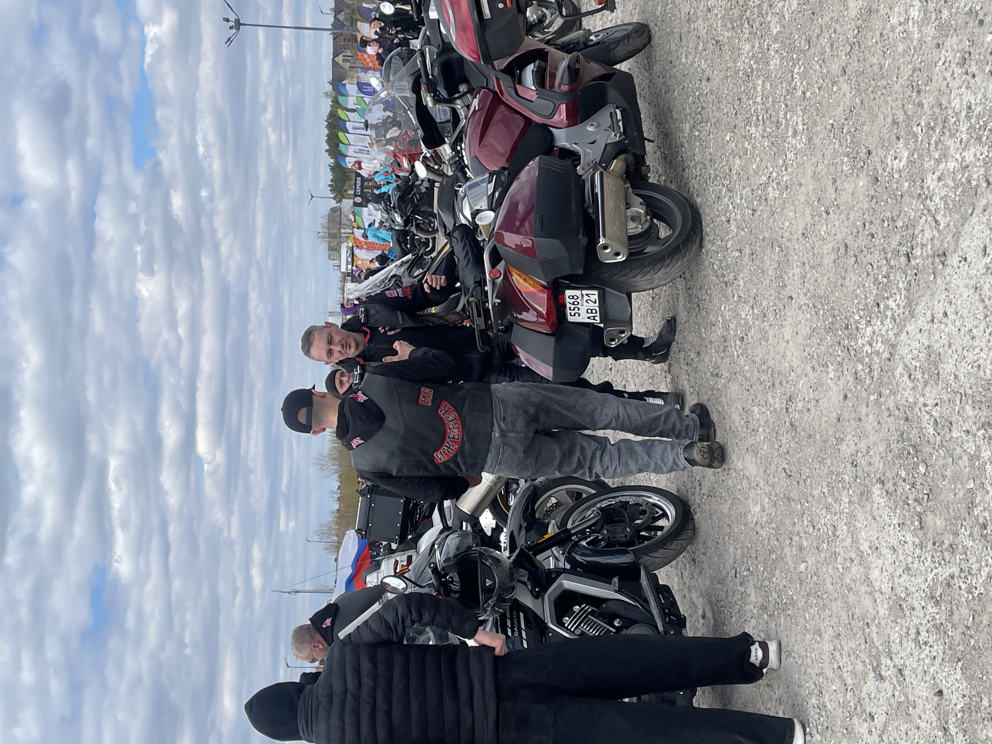 The image shows a group of people standing around motorcycles in an outdoor setting, likely a parking area or open space, in Naberezhnye Chelny, Russia. Several motorcycles are parked close together. Some individuals are wearing vests with patches, possibly indicating a motorcycle club affiliation. One motorcycle has a license plate with Russian text: "5568 AB 21". In the background, there are flag-like banners, with what appears to be event marketing/advertising text on them. The sky overhead has a cloud cover. The location is a gravel/asphalt surface, potentially a parking lot. There are at least four people visible, with one individual in the center making a hand gesture. There are multiple additional motorcycles with varying sizes.