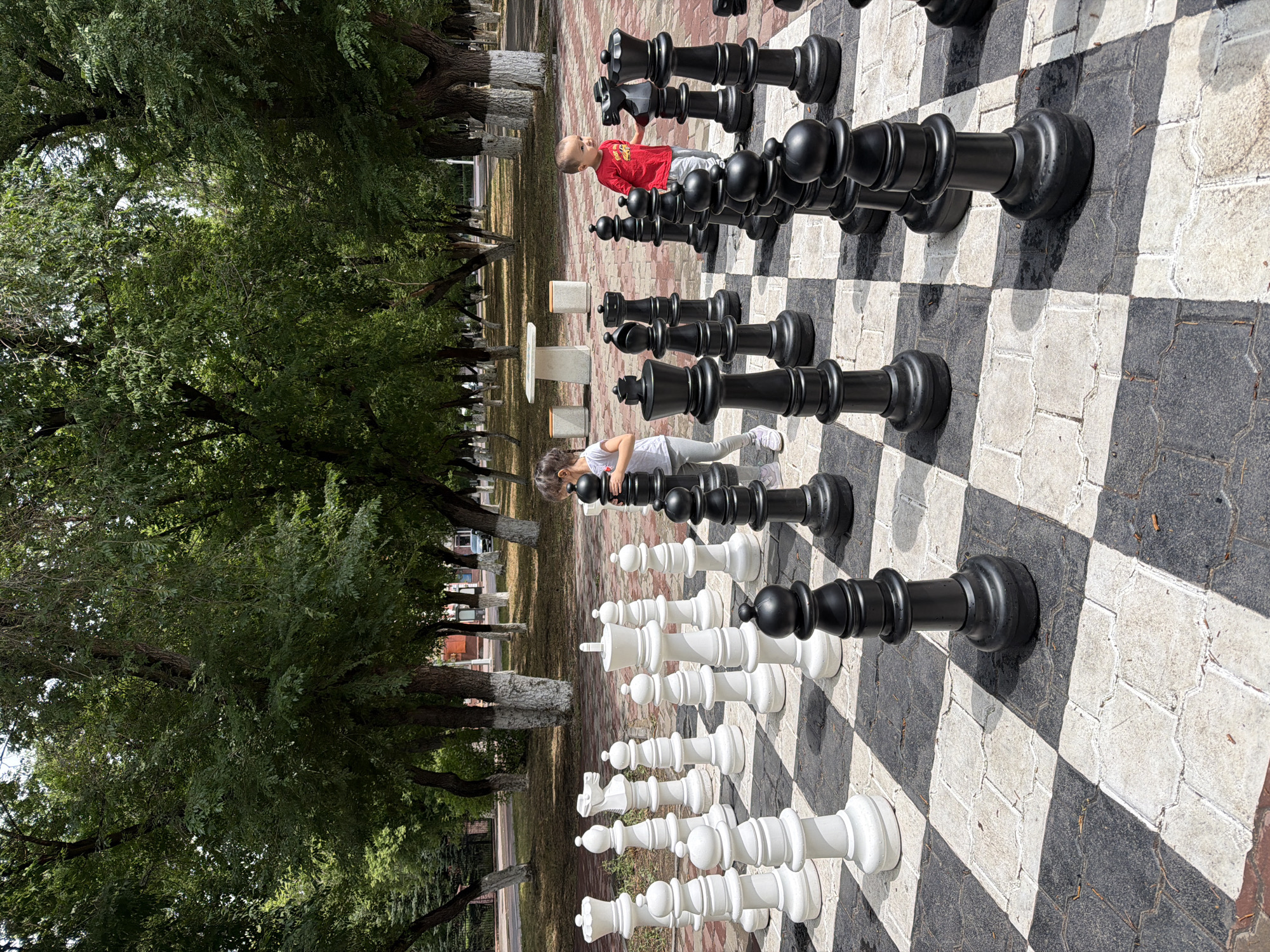 The image depicts an outdoor giant chessboard in a park setting in Karaganda, Kazakhstan. Two young children are interacting with the chess pieces. A girl in a grey t-shirt and leggings stands on the left side of the board, near a white pawn and a white knight. A toddler in a red t-shirt and grey pants is on the right side, positioned between black pawns and a black knight. The chessboard is laid out on a paved area, and the surrounding environment features mature trees with dappled sunlight, suggesting a daytime scene in a park or public square. Several white chess pieces, including pawns, a knight, and a king, are visible on one side of the board, while black pieces, including pawns, a bishop, and a king, are on the other. The background shows more trees, park benches, and a table, indicating a recreational area within the city.