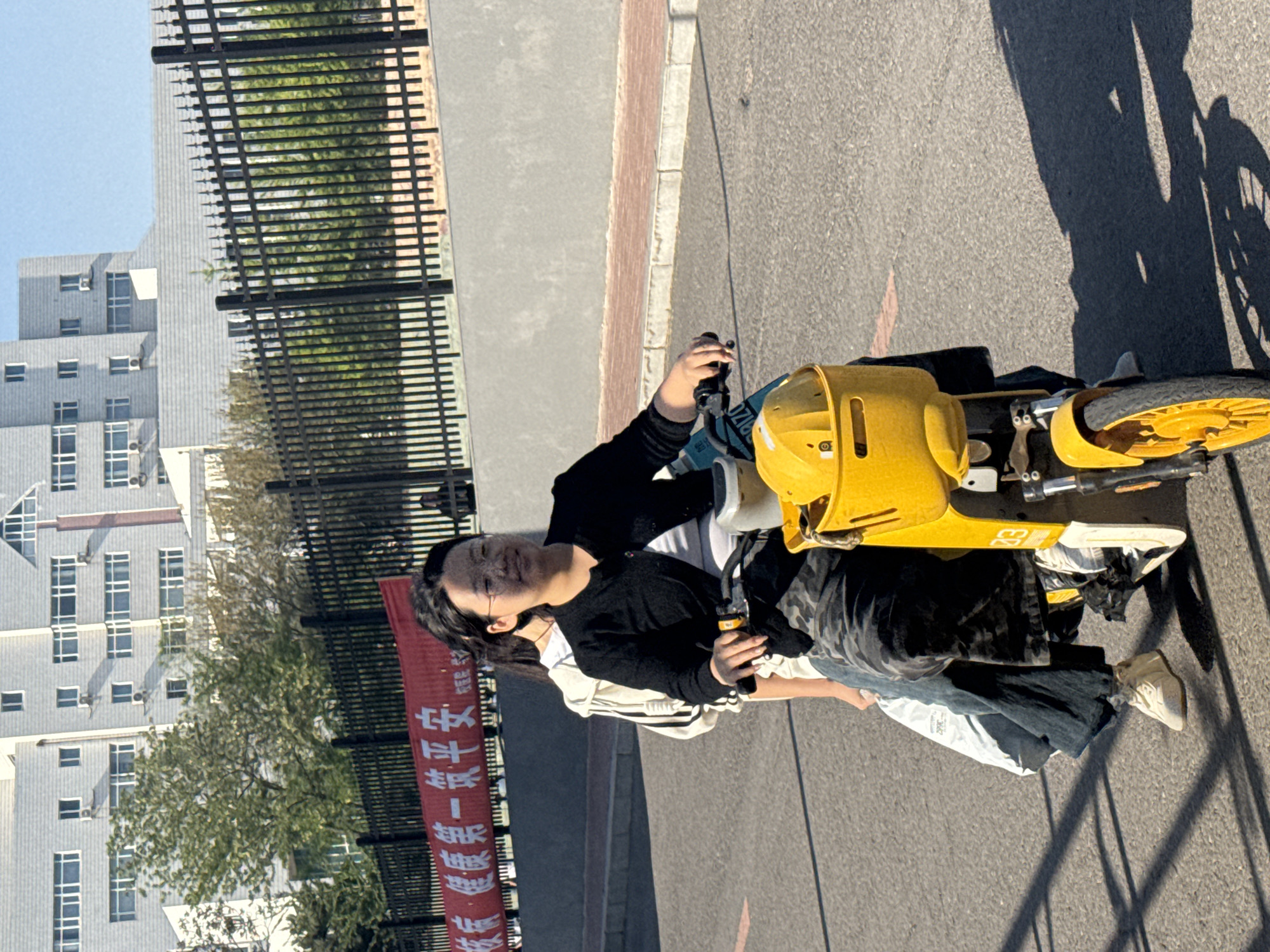 A horizontal medium shot shows a young woman in Zhongtun, China, seated on a yellow electric bicycle, appearing to operate its controls. She is wearing a black cardigan over a white top, and jeans, with white sneakers on her feet. Her hair is dark, and she wears glasses. The bicycle is positioned on an asphalt road. The shadow of the bicycle is visible on the right side of the frame. A black metal fence runs along the left side of the road. Behind the fence, a red banner with Chinese characters is displayed. Trees and a multi-story building are visible in the background. The sky is clear and light blue.