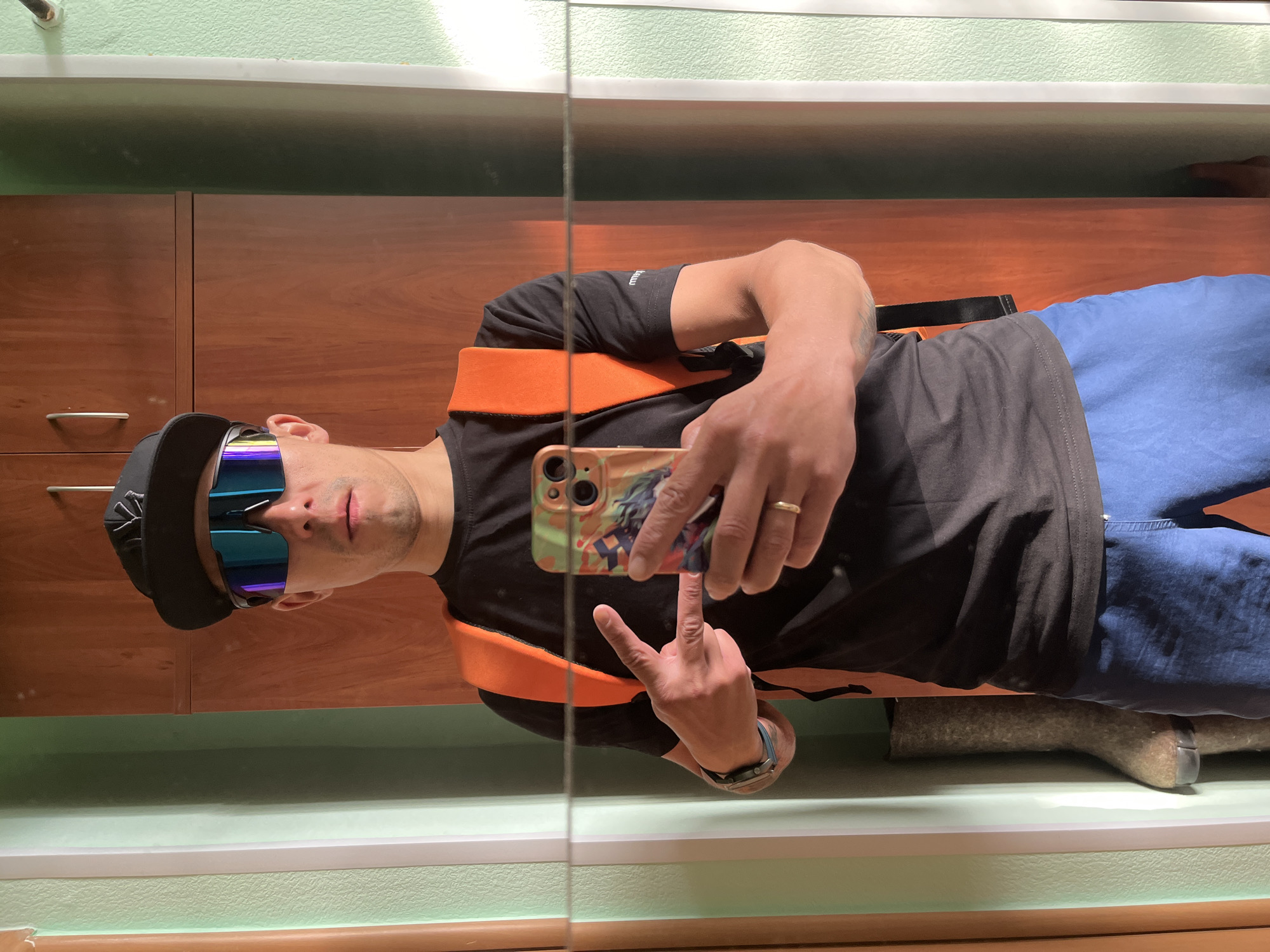 A man is reflected in a mirror. He is wearing a black baseball cap with a white logo, blue sunglasses, a black t-shirt, blue jeans, and an orange harness. He is holding a cell phone, taking a selfie, and giving a peace sign. There is a wedding band on his left ring finger and a watch on his left wrist. The background consists of wooden cabinets, a light green wall, and a white trim. Behind him is a grey valenki (traditional Russian felt boot). The location is likely an interior space in Chaikovsky, Russia.