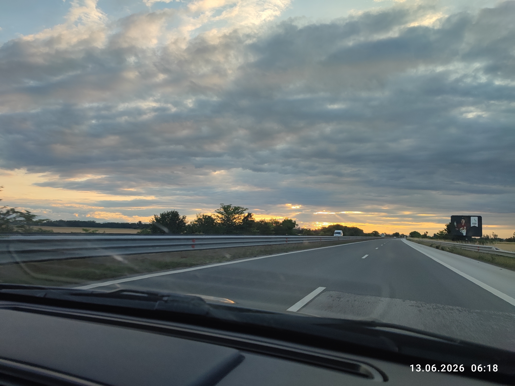 The image captures a view from inside a vehicle traveling on a highway, likely on the outskirts of Tsaratsovo, Bulgaria. The perspective is from the driver's seat, looking forward through the windshield, which shows some dust and wiper streaks. The highway stretches ahead, with multiple lanes marked by white lines. Guardrails are visible on the left side of the road.
The sky is filled with dramatic, cloudy formations, with patches of sunlight breaking through, suggesting either sunrise or sunset. The warm hues of the setting or rising sun are visible on the horizon, casting a golden glow. A billboard is present on the right side of the highway, featuring an advertisement with an image of a person. In the distance, several vehicles, including a white van, are driving on the highway. The landscape flanking the road appears to be rural, with fields and trees lining the horizon. The timestamp "13.06.2026 06:18" is overlaid on the bottom right corner of the image.