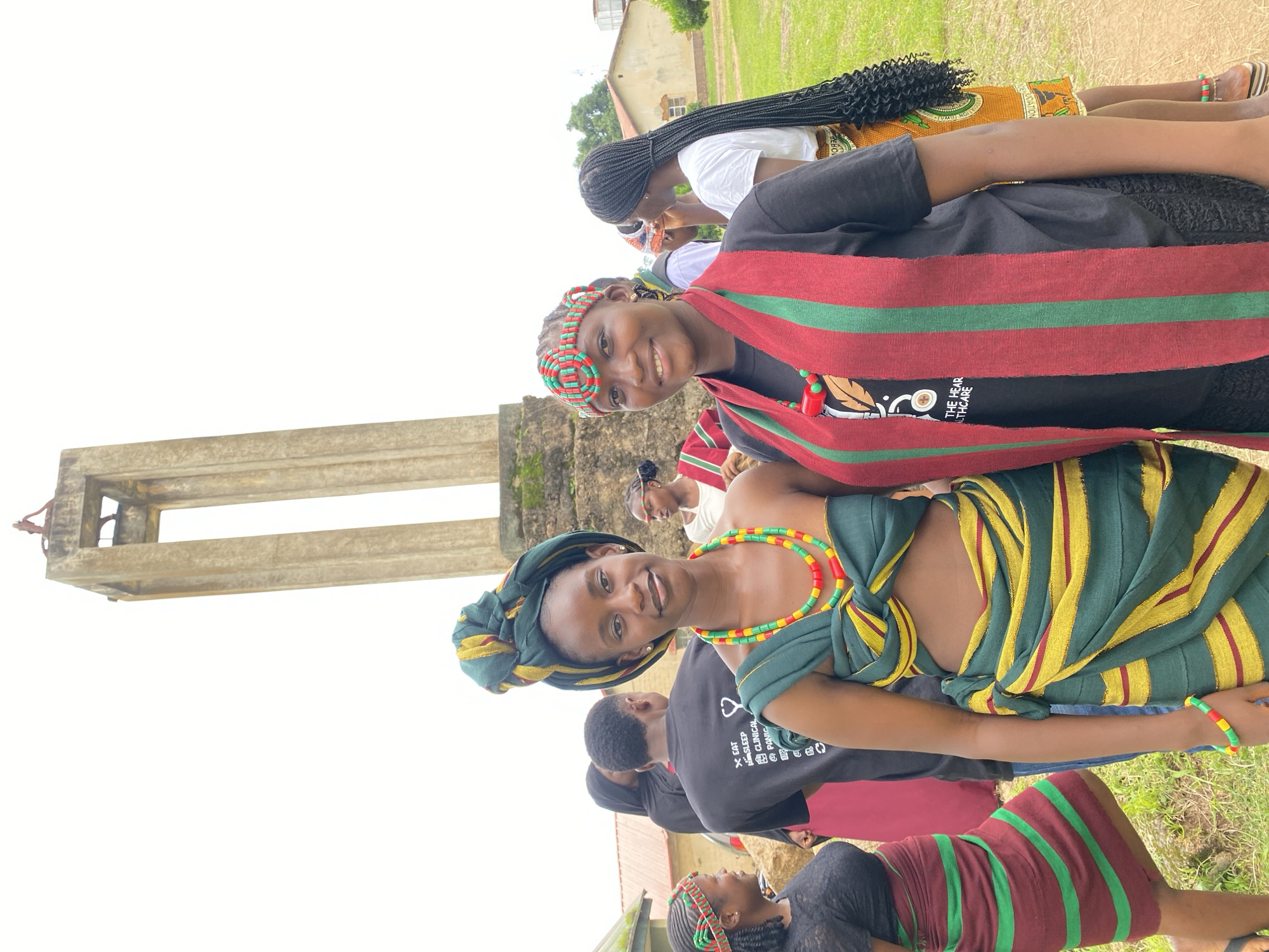 In Wang, Nigeria, a group of young people are gathered outdoors, possibly for a cultural event or celebration. Two young women are prominently featured in the foreground, both smiling and looking towards the camera. They are dressed in vibrant, traditional attire.
The woman in the front left wears a green and yellow striped wrap over a black t-shirt, adorned with a colorful beaded necklace and matching headband. Her hair is styled in a traditional wrap. The woman behind her, slightly to the right, wears a red and green striped sash over a black t-shirt, with a similar beaded headband and necklace. Her hair is in long, dark braids. Another young woman in the background, also in traditional attire, is partially visible.
The setting appears to be a grassy area with a concrete structure, possibly a pillar or monument, on the left side of the frame. A building with an orange roof and windows can be seen in the background, suggesting an outdoor gathering. The sky is overcast, indicating a cloudy day. The overall mood is festive and joyful, with the subjects appearing relaxed and happy. Some of the visible text on the t-shirt of the woman in the front right reads: "EAT SLEEP CLINICAL @ PANIC".