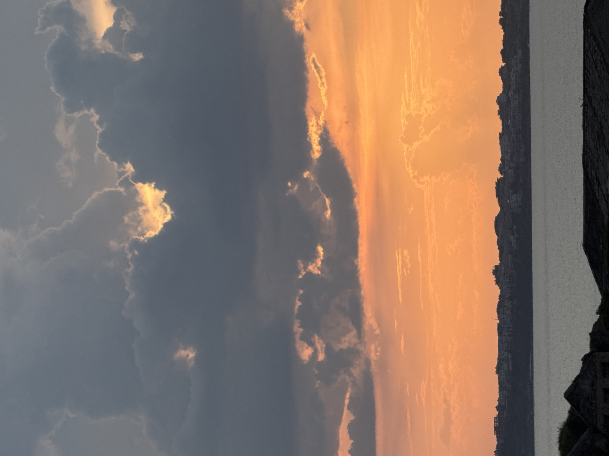 The image captures a striking sunset or sunrise over the sea, likely off the coast of Sozopol, Bulgaria. The sky is dominated by a dramatic contrast of colors and cloud formations. On the right side, the sky glows with intense orange and yellow hues, indicative of the sun's low position, illuminating various wispy and textured clouds from behind. These clouds range from soft, diffused orange to brighter, glowing streaks along their edges.
Towards the left, the sky transitions into a much darker, greyish-blue, filled with heavy, dense cloud cover. Despite the darkness, pockets of brilliant golden light pierce through or reflect off these clouds, creating intricate patterns and highlights.
Below the expansive sky, a calm body of water, likely the Black Sea, stretches horizontally, reflecting the subdued light. Along the distant horizon, a dark silhouette of a coastline is visible, featuring numerous structures and buildings that suggest a coastal town or city. In the far right foreground, dark, rugged landmasses or rocks frame the scene, adding depth. The overall scene conveys a sense of dramatic natural beauty and tranquility at dawn or dusk. There is no visible text in the image.