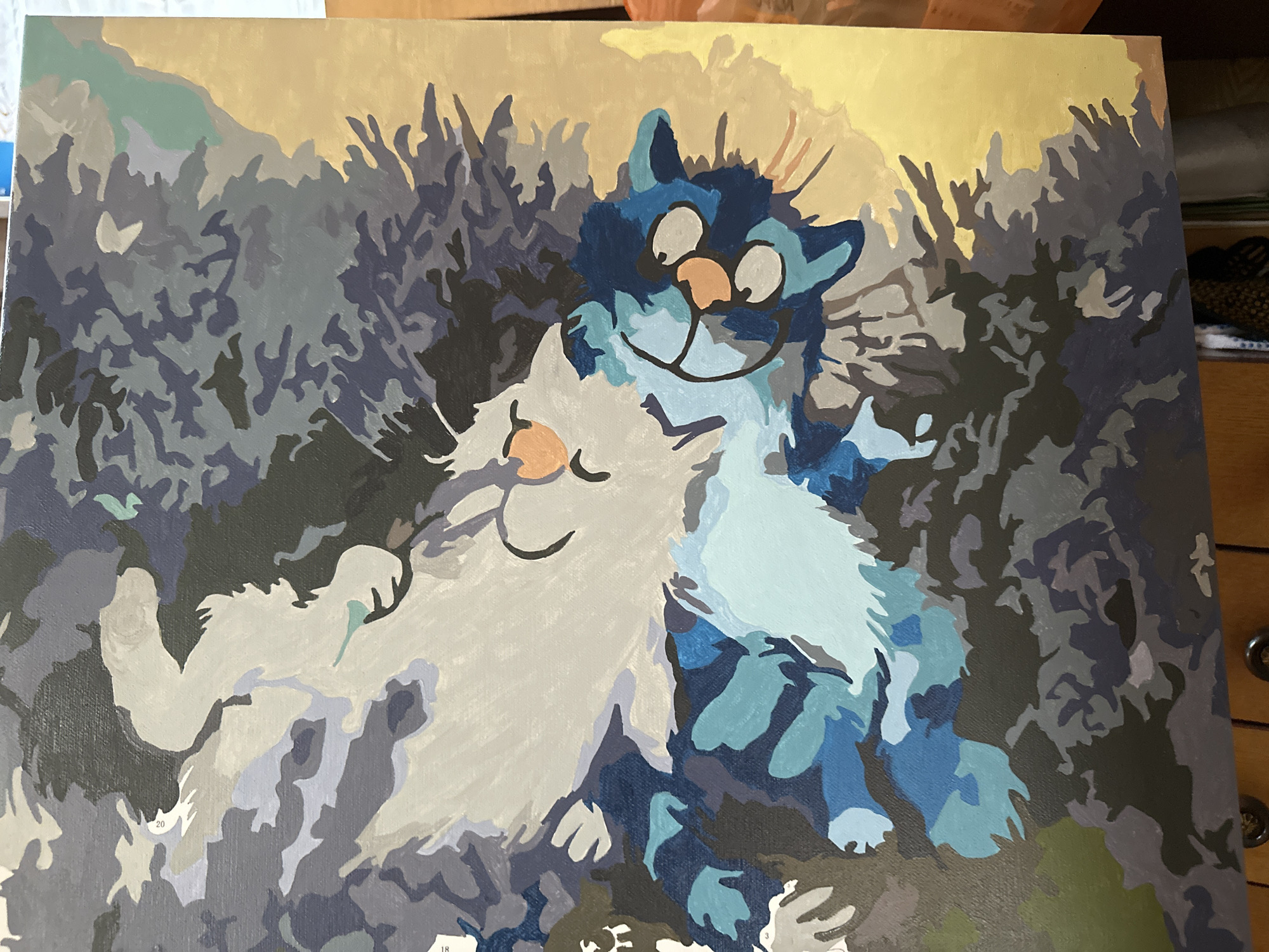 A painting depicts two stylized cats against a background of abstract foliage and a light yellow sky. One cat is predominantly white and gray, with a pink nose and a closed eye, suggesting it is sleeping or relaxed. The other cat is primarily blue, with lighter blue patches and a dark blue head. It has a pink nose, large white eyes with black pupils, and a smiling mouth. The background consists of various shades of gray, blue, and black, suggesting dense vegetation or foliage. The overall style is cartoonish and whimsical. The location in the city of Среднее Кечево, Russia is not discernible from the painting.