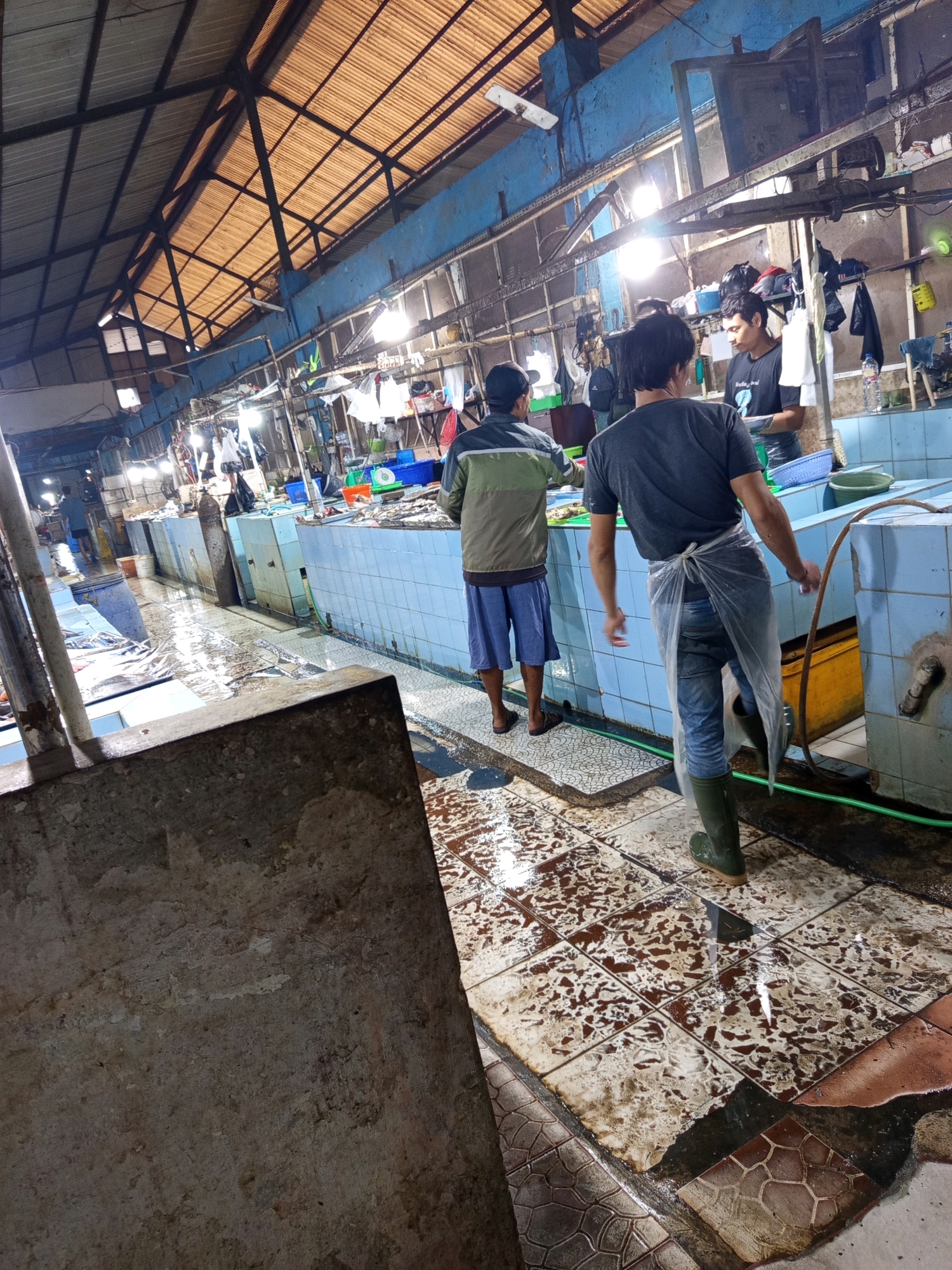 The image depicts an indoor market scene, likely a fish market, in Karawang, Indonesia. In the foreground, a large, rough concrete structure occupies the left side of the frame. The floor throughout the market is wet and tiled, with some tiles appearing stained or worn. The stalls are lined with blue tiled countertops, typical of fish markets, and some contain visible fish.
Three men are present in the scene. One man, seen from the back, is wearing a grey t-shirt, blue jeans, and green rubber boots, with a transparent plastic apron tied around his waist. He appears to be standing near a stall. Another man, also seen from the back, is wearing a green and grey jacket and blue shorts. A third man, wearing a black t-shirt with a graphic, is partially visible behind the man in the blue apron.
The market has a high, sloped roof constructed with wooden beams and corrugated roofing material, allowing natural light to filter through. Several bare light bulbs hang from the ceiling, illuminating the market. The overall environment appears to be a functional, working market, with evidence of recent activity and water on the floor. The presence of fish and the wet conditions are characteristic of a place where seafood is sold and processed.