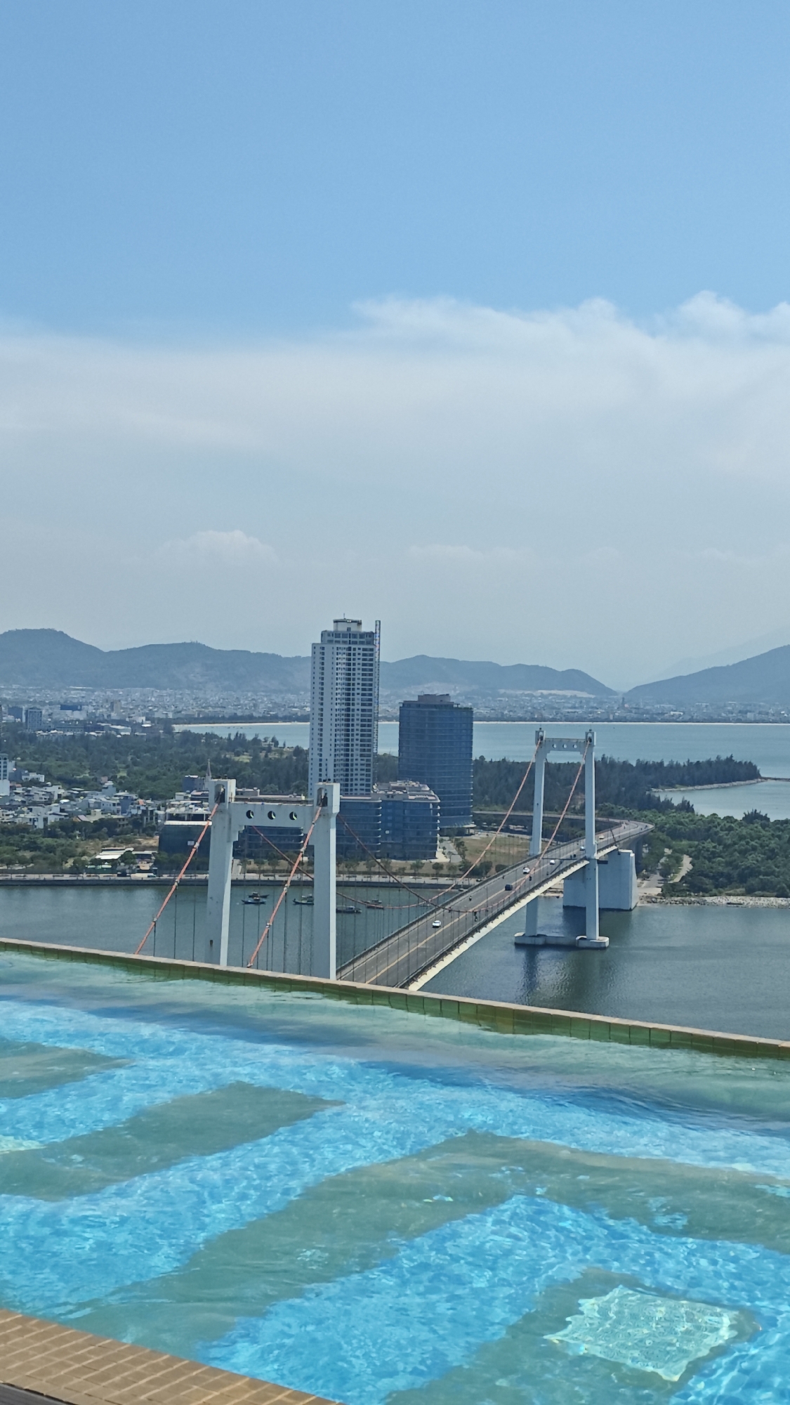 The image presents a high-angle view of the Tran Thi Ly Bridge in Đà Nẵng, Vietnam. The bridge, a cable-stayed structure, is seen spanning a body of water, likely the Han River. Cars are visible on the bridge, indicating it is in use. In the background, the city skyline is visible, including buildings and mountains in the distance under a blue sky with some clouds. In the foreground, the edge of a swimming pool is seen, with blue tiles and water visible, suggesting the photo was taken from a high-rise building or hotel.