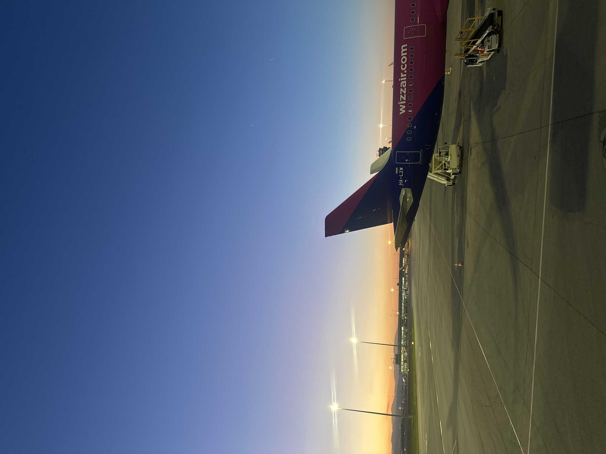 The image shows the tail of a Wizzair airplane at an airport apron at dusk. The plane is registered as HA-LZW. A passenger boarding bridge and other ground support equipment are near the plane. The apron is marked with painted lines. The sky transitions from blue to orange in the distance. Airport lighting is visible. In the far distance, buildings and mountains are silhouetted against the horizon. Given Wizzair's operating base and the registration of HA-LZW, it is possible this is in Sofia, Bulgaria.
