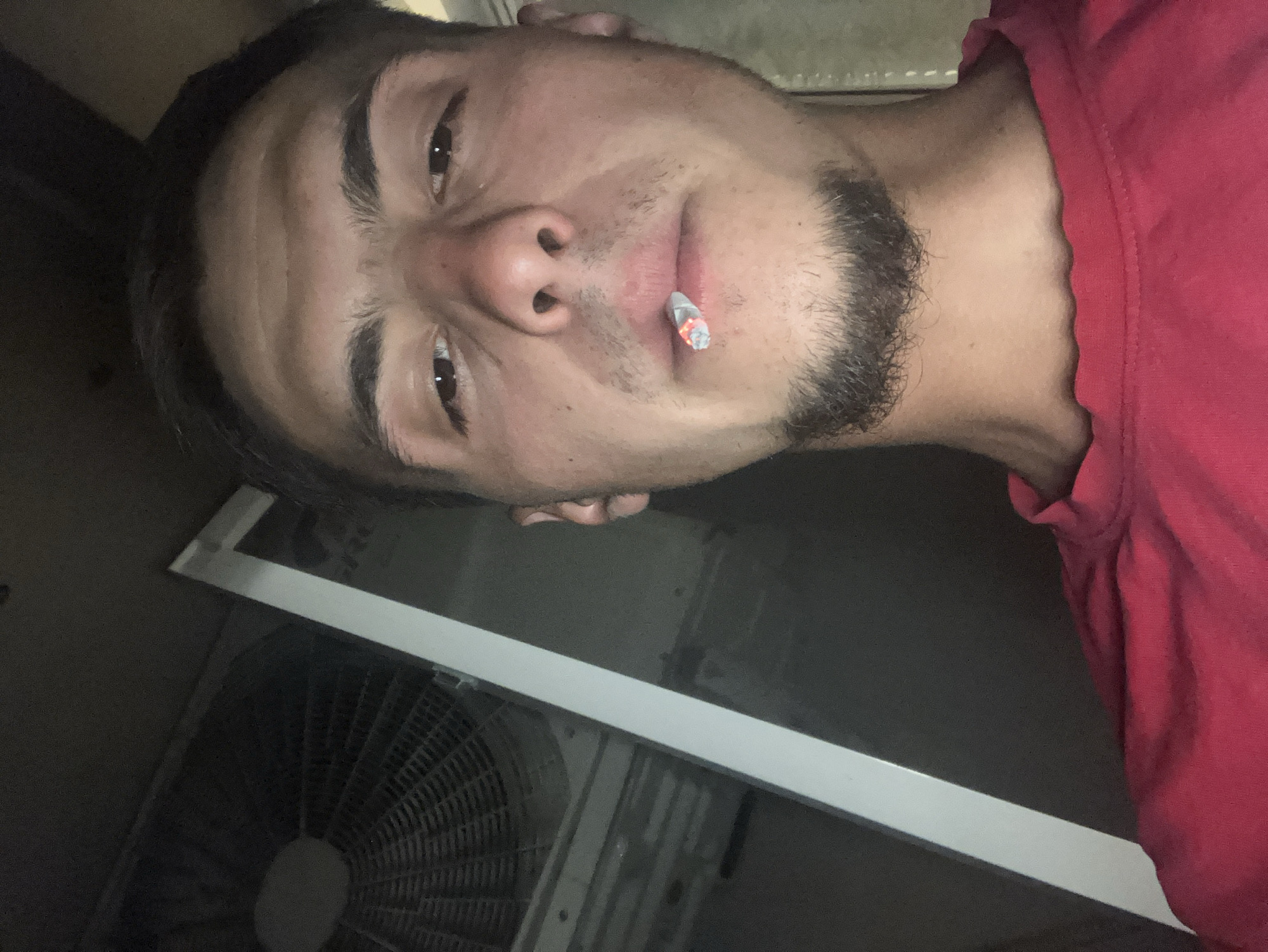 A close-up, angled shot depicts a man with a trimmed beard and dark hair, holding a lit cigarette between his lips. He is wearing a red shirt. In the background, a window air conditioning unit is visible. The location of the scene in Plovdiv, Bulgaria cannot be determined from the visual information provided.