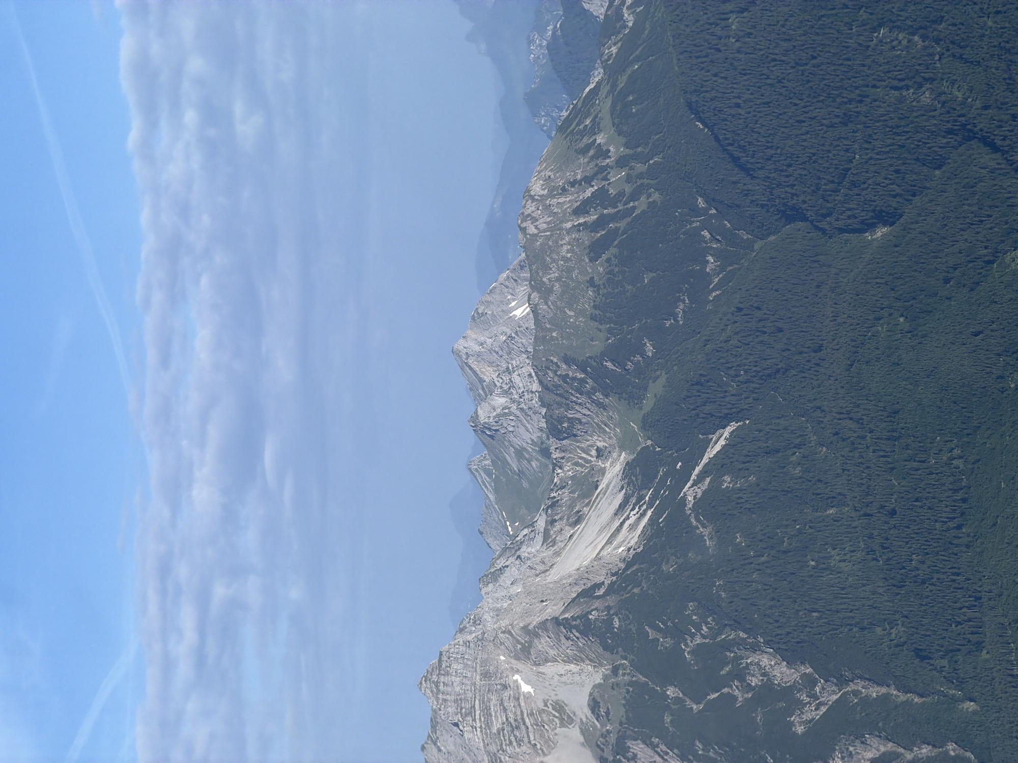 This is a landscape photograph taken from a high vantage point, looking out over a mountain range. The sky is a clear blue with some wispy clouds and a faint contrail. The mountains are rugged and imposing, with rocky peaks and steep slopes. The lower slopes are covered in dense green forest, which appears to be coniferous. There are patches of snow on some of the higher, shaded areas of the mountains. The overall impression is one of natural grandeur and remoteness. The specific location in Grainau, Germany is a mountain viewpoint offering panoramic views of the Alps. There are no people or man-made structures visible in this scene.