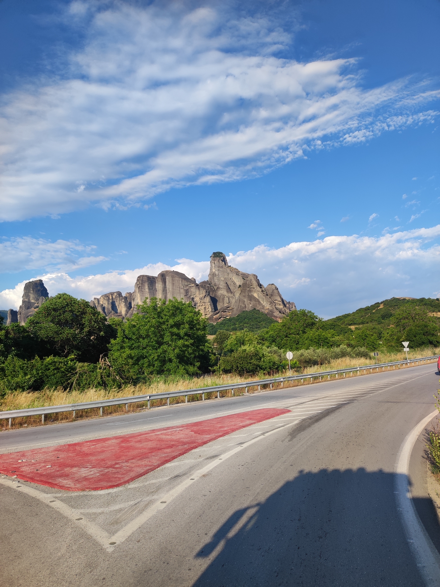 The image depicts a scenic view of the Meteora rock formations near Kalabaka, Greece. The foreground features a two-lane asphalt road with a distinct red-painted area on the left side, possibly indicating a pedestrian crossing or a special zone. White lane markings are visible on the road, and a silver guardrail runs along the side. Beyond the road, a dense line of green trees and vegetation stretches across the landscape, with dry grasses and bushes at the base of the trees. The backdrop is dominated by the imposing, weathered rock formations of Meteora, with their unique geological shapes and vertical cliffs. The sky above is a vibrant blue, dotted with scattered white clouds. A shadow of what appears to be a vehicle is cast on the road in the lower right portion of the image. The lighting suggests it is daytime, likely late afternoon given the long shadow. The overall scene conveys a sense of natural beauty and geological wonder.