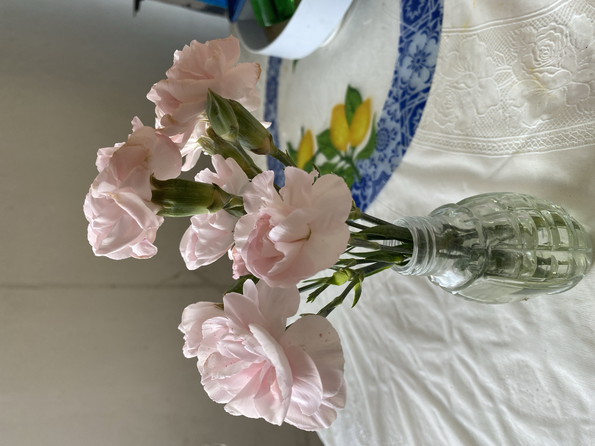 A close-up shot captures a small bouquet of pale pink carnations arranged in a clear glass vase. The vase, designed to resemble a grenade, sits on a white tablecloth with embroidered floral patterns. In the background, a round plate with a blue and yellow pattern is visible, along with a white container and green objects. The lighting suggests an indoor setting with natural light. The image does not provide any information about the city of Smolino, Russia.