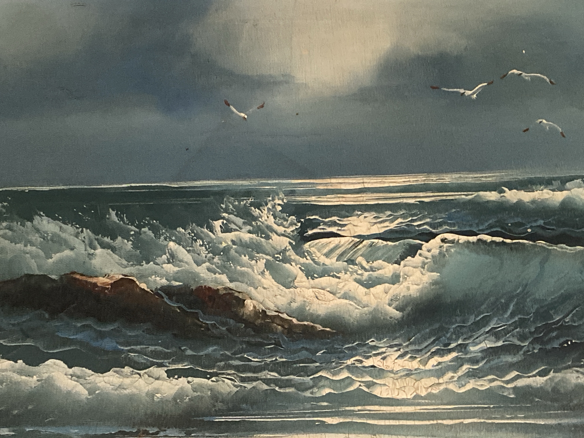 The image depicts a seascape painting, likely of an ocean scene. The foreground features rough, white-capped waves crashing against a rocky outcrop. The waves are rendered with energetic brushstrokes, emphasizing their dynamic motion. The rocks are dark brown and partially submerged. The background shows a dark, cloudy sky, possibly indicating an approaching storm. Above the waves, a few seabirds are scattered across the sky. The overall tone of the painting is dramatic and turbulent, with a strong contrast between light and shadow. There is no indication of it being in Tinaquillo, Venezuela.