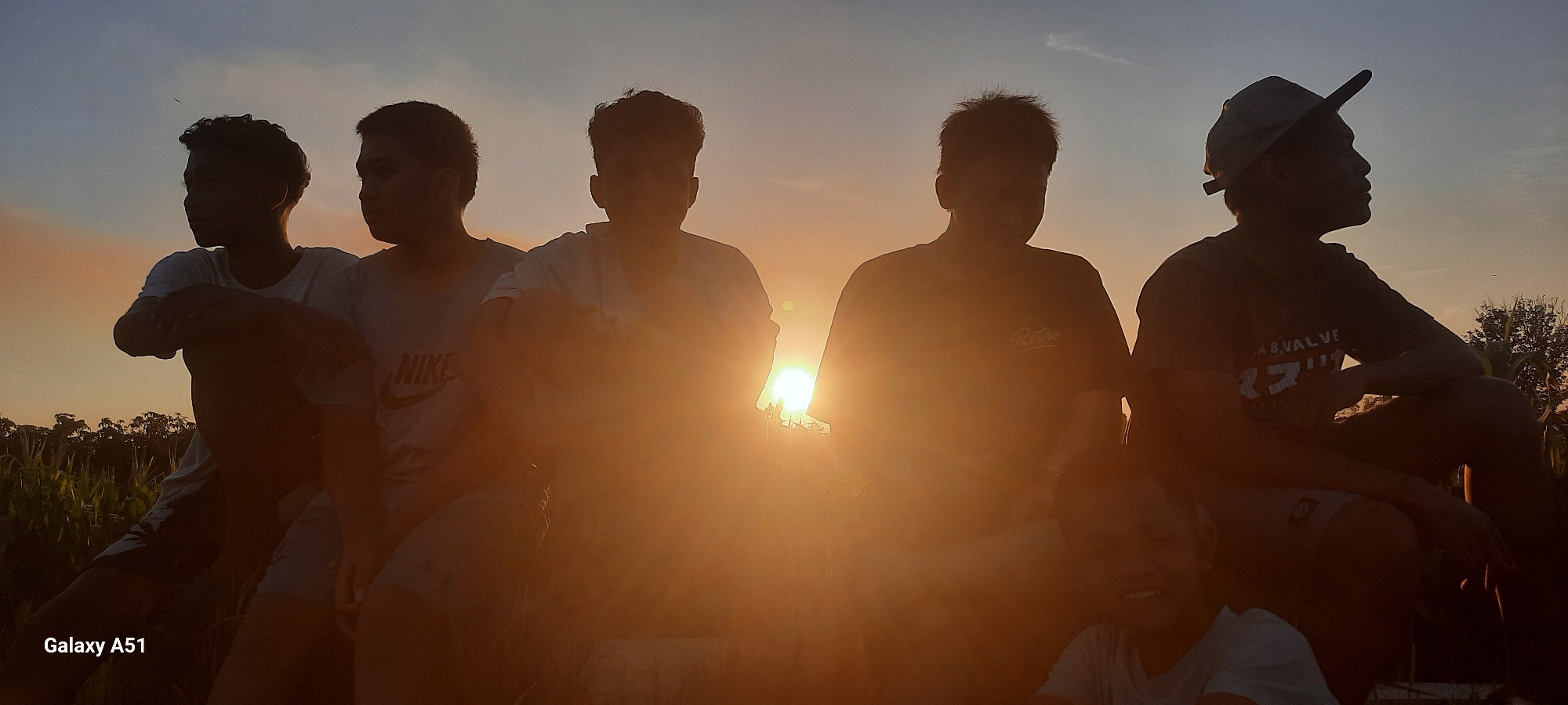 A group of six young males are silhouetted against a sunset in Duren, Indonesia. They are seated close together in a field, possibly a cornfield, with tall stalks visible in the foreground. The sky is a gradient of orange and blue, with the sun setting directly behind the central figures, creating a strong backlight.
The individuals, from left to right, are:
1. A young man with short, curly hair, wearing a t-shirt. He is looking to his left.
2. A young man with short, dark hair, wearing a t-shirt. He is looking to his right.
3. A young man with short, spiky hair, wearing a t-shirt. He is looking forward.
4. A young man with short hair, wearing a dark t-shirt with a graphic print. He is looking forward.
5. A young man wearing a baseball cap backward, looking to his right. His t-shirt has text on it, part of which reads "AB.VALVE".
6. A young male in the front, partially obscured, with short hair and wearing a t-shirt. He is smiling and looking towards the camera.
The overall mood is relaxed and convivial. The "Galaxy A51" watermark is present in the bottom left corner of the image, indicating the device used for capture. The setting appears to be rural, given the agricultural field and the natural light conditions of a sunset.