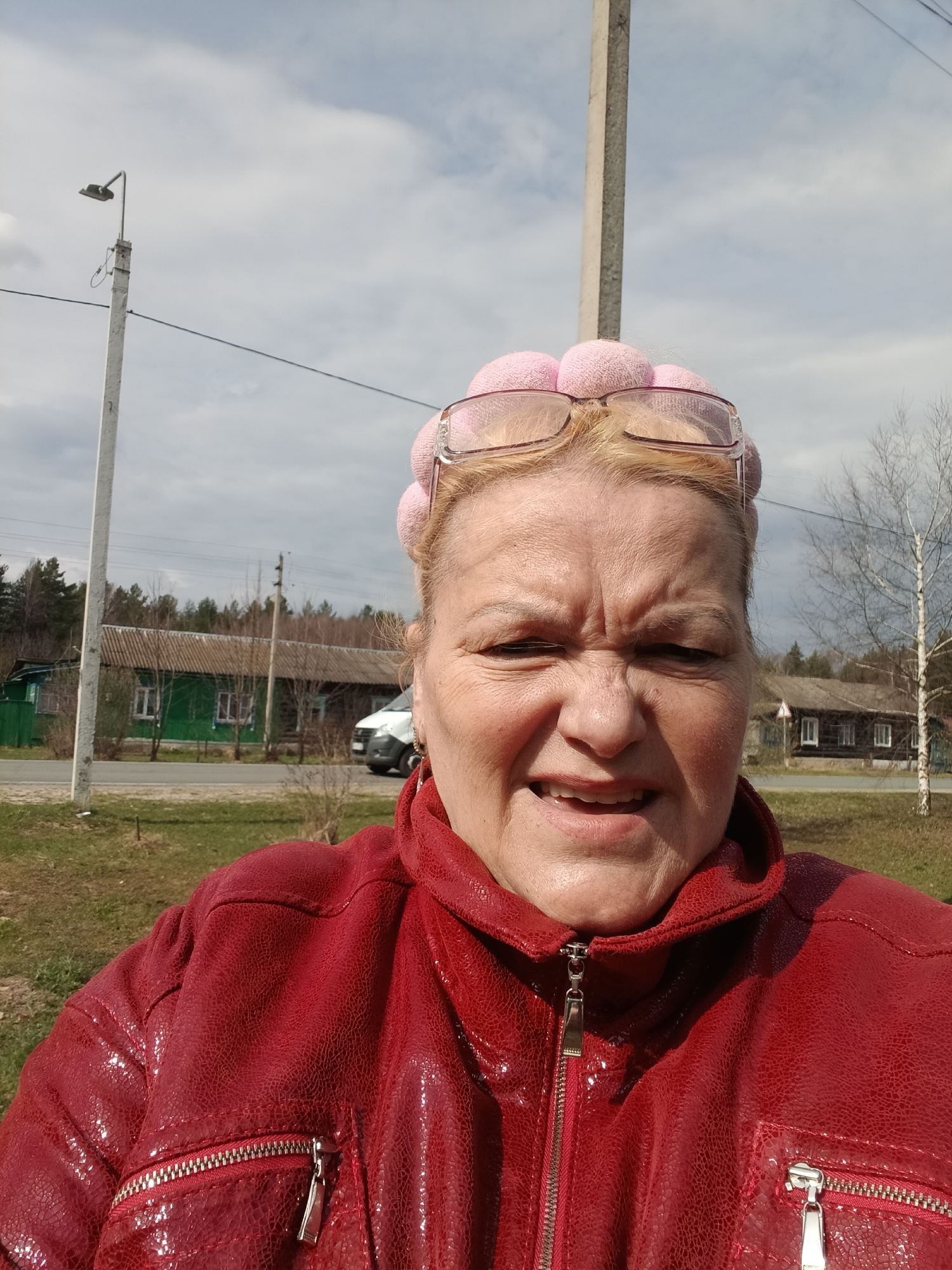A medium close-up selfie captures an adult woman, likely middle-aged, outdoors in a rural or semi-rural setting under a bright, cloudy sky. The woman has blonde hair and is wearing a distinctive pink, possibly knitted or fabric, headband with spherical elements, resembling large hair rollers or decorative pom-poms. Pink-framed glasses are pushed up onto her forehead, resting on the headwear. She is dressed in a shiny, textured red jacket featuring multiple zippers.
Her facial expression is prominent, characterized by a furrowed brow, wrinkled forehead, and squinted eyes, suggesting she might be grimacing, squinting against the light, or making a playful face. Her mouth is slightly open, revealing some teeth.
The background reveals a roadside scene on what appears to be a cool, early spring or late autumn day. To the left, a tall utility pole with a street lamp and power lines stands next to a patch of green grass. Further back, a long, green-painted wooden house with white window frames is visible, situated near a paved road where a white van is partially seen. A line of trees and a distant forest complete the left side of the background. On the right, another utility pole, a bare-branched birch tree, and a dark wooden house with white window frames are present. The ground is covered with sparse grass and some bare earth. No legible text is visible in the image. The setting suggests a village or small settlement, consistent with the location hint of Красный Маяк, Russia.