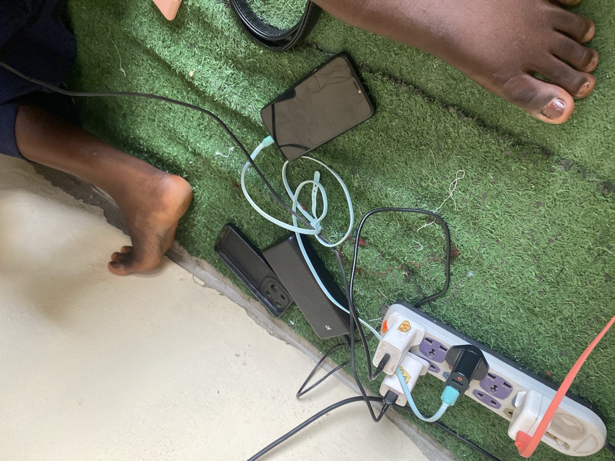 In Jauro Yinu, Nigeria, a scene unfolds on a patch of artificial grass adjacent to a cream-colored wall. The focus is on a tangle of electronics and charging cables. A white power strip, plugged into an unseen source, has several devices connected. A black phone is charging via a light blue cable, while another black device, possibly a power bank, is also connected. Two black phone cases are stacked near the wall, one on top of the other.
The lower right portion of the image reveals the feet of two individuals, positioned close to the electronic setup. One person's feet are mostly within the frame, with dark skin and light-colored toenails, resting on the artificial turf. Another person's bare foot is visible in the upper right quadrant, positioned near the wall. The overall environment appears to be indoors or in a covered outdoor space, indicated by the wall and the presence of electrical outlets. The arrangement of the charging devices suggests an active attempt to power multiple electronic items simultaneously.