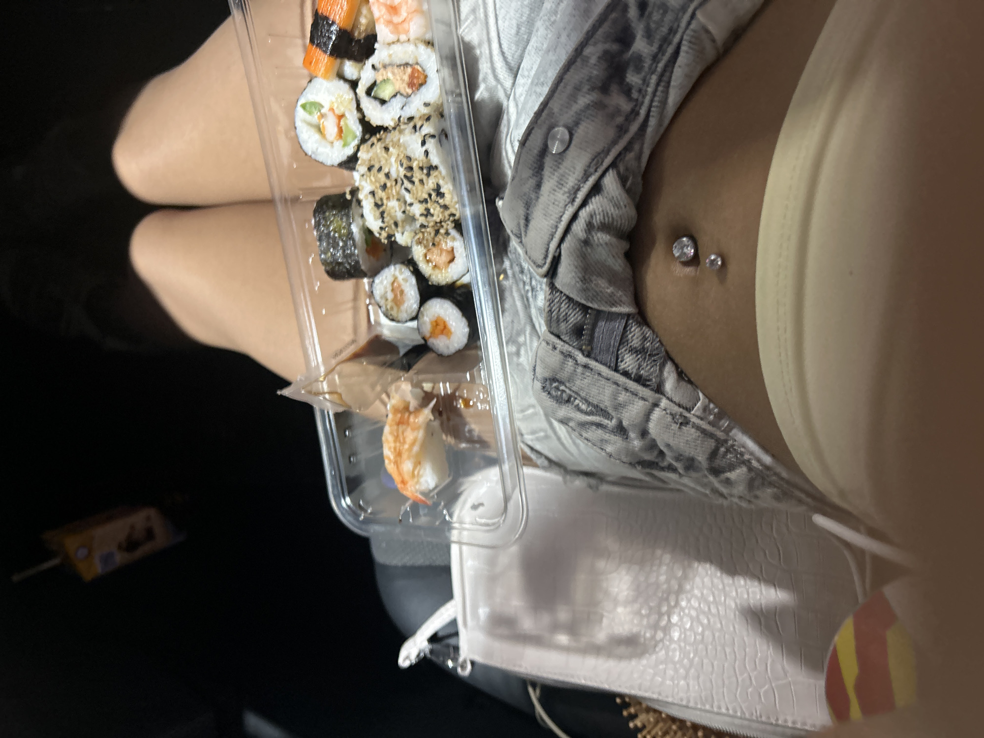 In Plovdiv, Bulgaria, a close-up shot shows a person's midsection holding a plastic container of sushi. The container holds various types of sushi rolls, some with sesame seeds and others with visible ingredients like shrimp and cucumber. To the left of the sushi, a white purse with a textured surface is visible. The person is wearing denim shorts and a light-colored top, with a belly button piercing. A small, brown carton with a straw is visible in the upper left corner of the frame. The background is dark and out of focus, suggesting an indoor or nighttime setting.