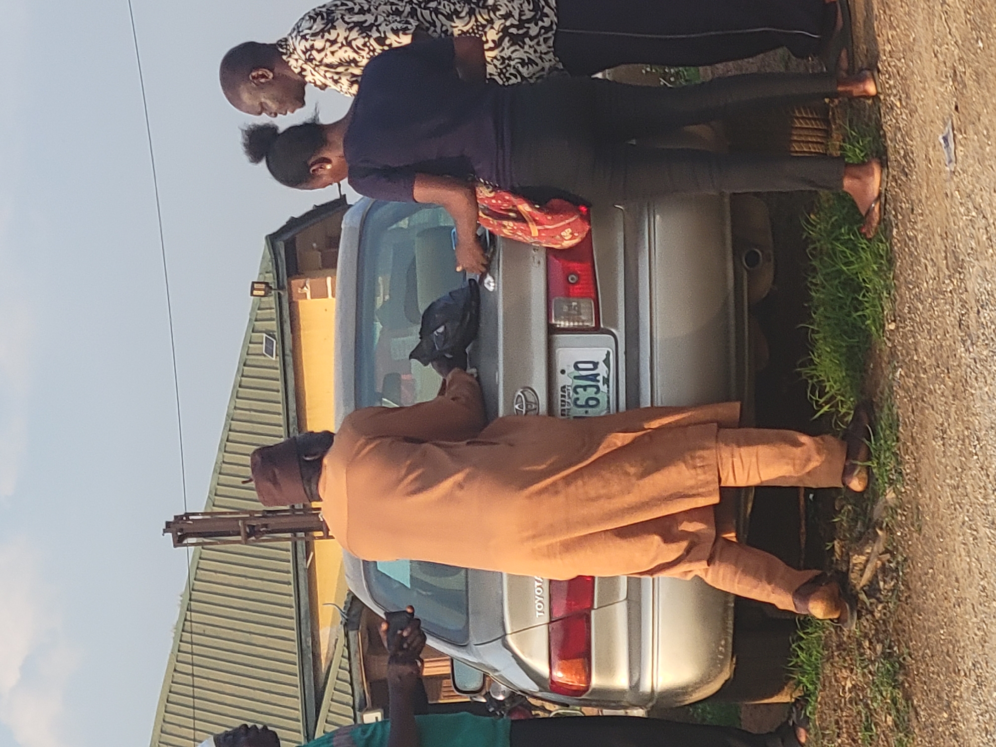In Kwamba, Nigeria, a group of people stand near the trunk of a silver Toyota car. A man in a brown traditional robe and cap is positioned at the trunk, appearing to interact with it. Two other people, a man in a patterned shirt and a woman in a blue shirt and a red bag, are also near the trunk. The car's license plate, "NYIA 63AQ", is visible. There is green grass along the roadside, and a building with a green roof is in the background.