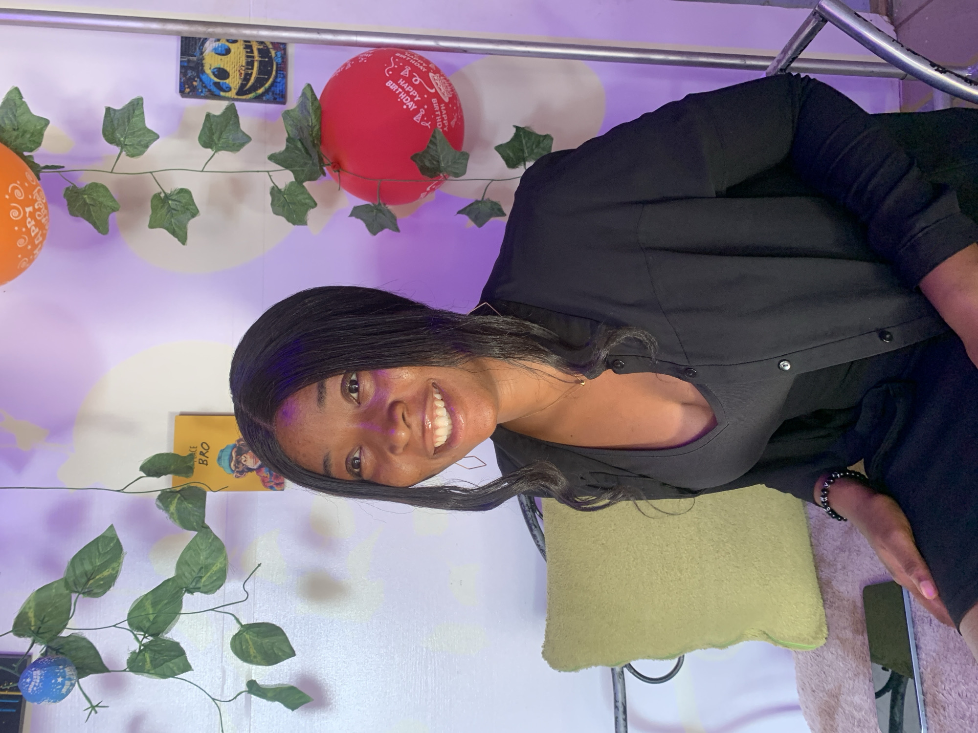 A woman is in a room in Jos, Nigeria. The woman is light-skinned with long black hair, wearing a black top. She is smiling. Decorations in the room include balloons, artificial vines, and a painted image on the wall. There is a light-colored wall with white cloud-like shapes painted on it. A small yellow book is also visible. A green cushion is nearby.