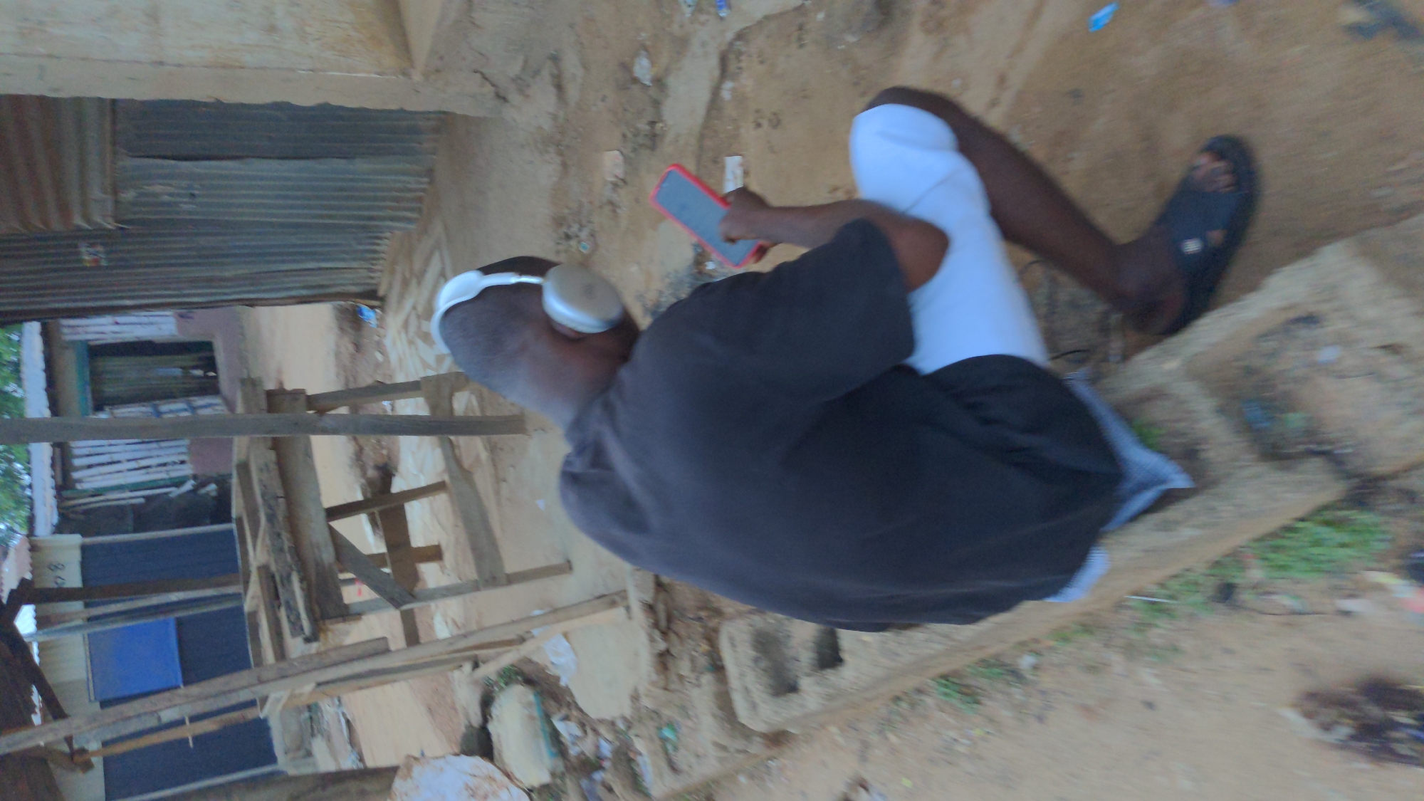 A person is seated outside near a building constructed of corrugated metal and lumber. The individual is wearing white pants, a black shirt, and a black sandal on one foot. They have headphones on and are holding a red phone. The surrounding environment appears to be a sandy area. The location is Masaka, Nigeria.