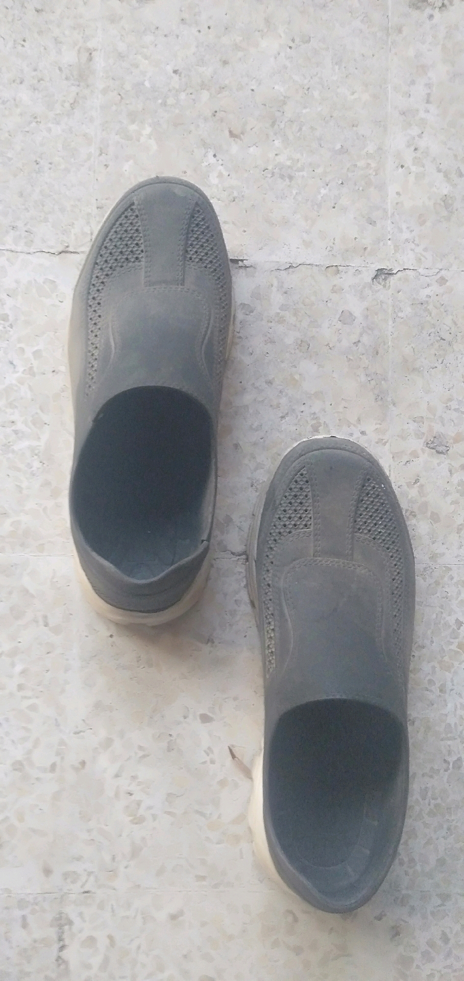 A pair of dark gray slip-on shoes are positioned on a light-colored tiled floor. The shoes are of a casual design, possibly made of a breathable material given the perforated pattern on the upper part. One shoe is positioned slightly higher and to the left of the other, with its opening facing upwards. The floor appears to be made of terrazzo tiles, with visible grout lines and some minor cracks or imperfections. The overall scene suggests an indoor or outdoor area with a tiled surface, common in residential or public spaces. There is no indication of any specific location within Plemahan, Indonesia, based solely on the visual information provided in the image.