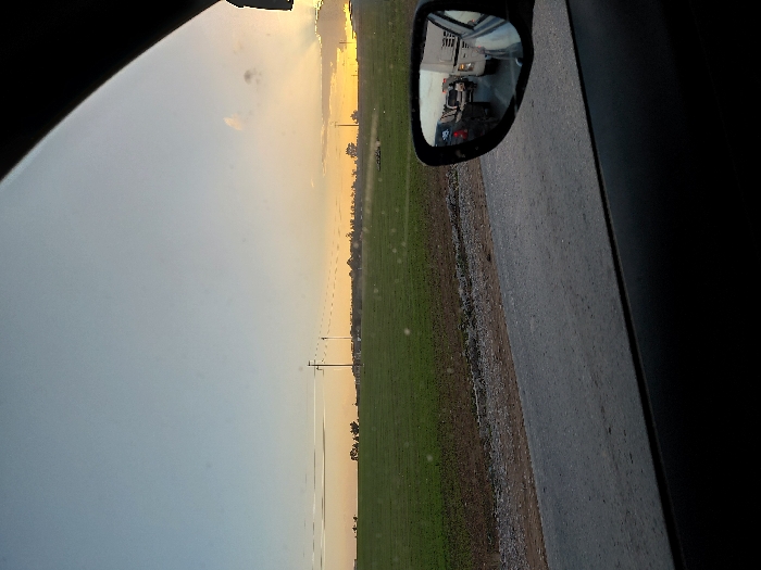 A car ride view presents a landscape. A side mirror reflects a vehicle. Beside the car, a paved road separates a grassy field. The horizon shows a mix of sky and possibly a setting sun. Utility poles stand along the edge of the field. The scene suggests a rural setting, likely captured from a moving car.