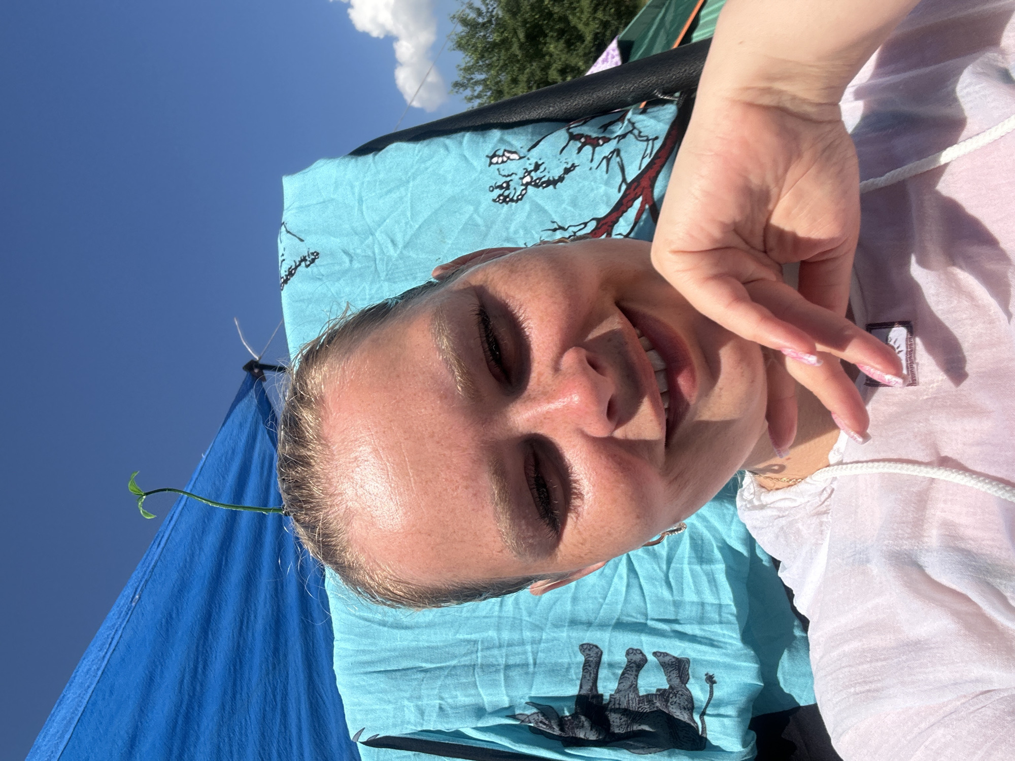 The image is a close-up, selfie-style shot of a young woman with blonde hair pulled back, facing the camera and smiling. She is outdoors on a sunny day, with a clear blue sky in the background. A blue tarp is visible behind her, along with a light blue towel featuring an elephant print. A small green sprout is growing from the top of her head, seemingly attached to something unseen. She is wearing a light-colored hooded shirt. Her right hand is raised, with her fingers gently touching her chin. The scene appears to be at a campsite or outdoor gathering, as indicated by the tents and natural surroundings in the background. The location is specified as the city of Жемчужный, Russia.