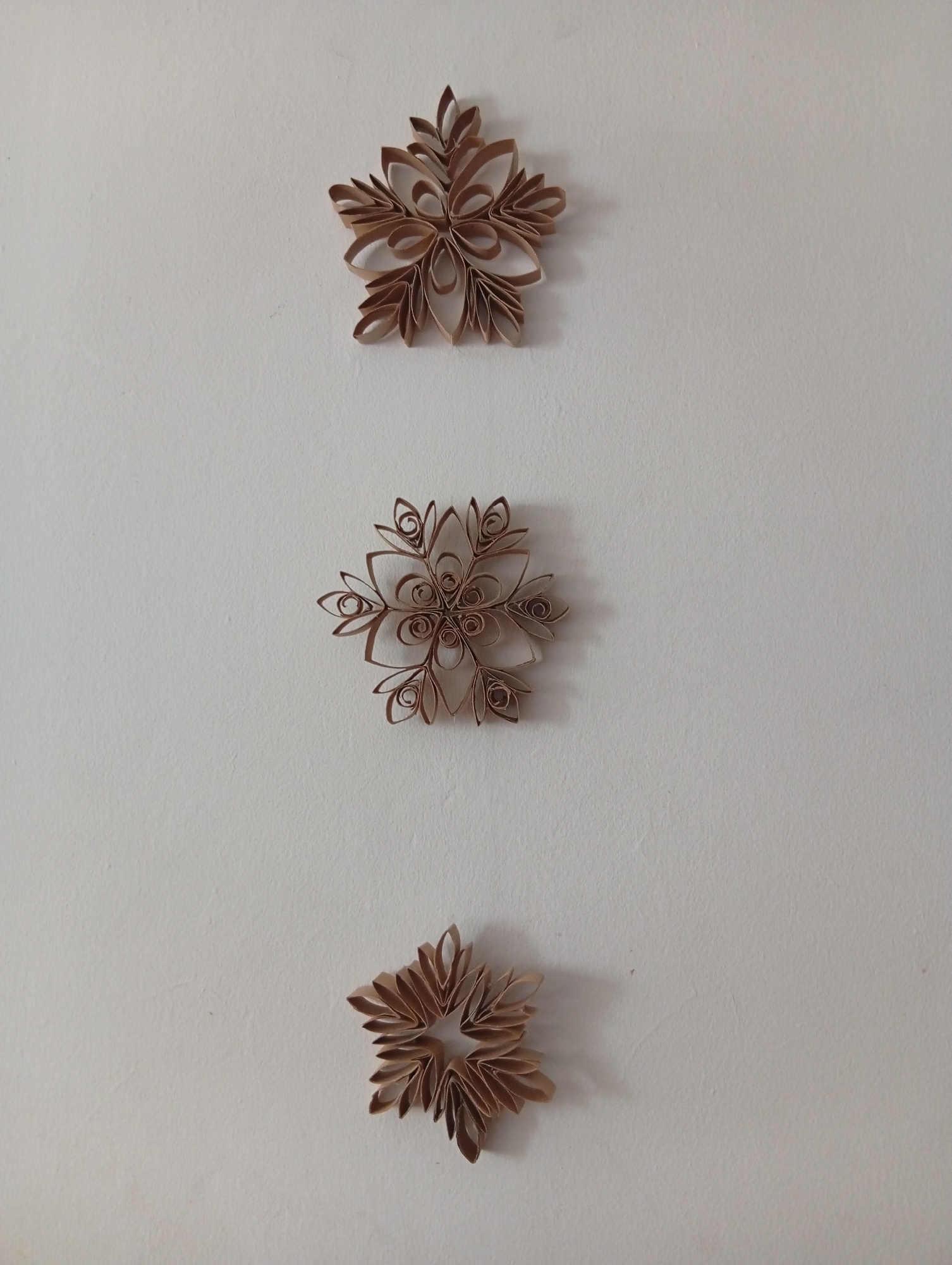 Three snowflake-shaped ornaments, crafted from what appear to be cut and curled toilet paper rolls, are affixed to a plain, off-white wall. The ornaments are arranged vertically, one above the other, with the top ornament positioned in the upper third of the frame, the middle ornament in the center, and the bottom ornament in the lower third. Each snowflake has a symmetrical design with multiple points or petals extending outwards. The middle snowflake is the most intricate, featuring smaller coiled elements within its design. The lighting suggests a neutral indoor environment. The scene is devoid of people or any specific urban context, making it impossible to determine its location within San Antonio de Los Altos, Venezuela.