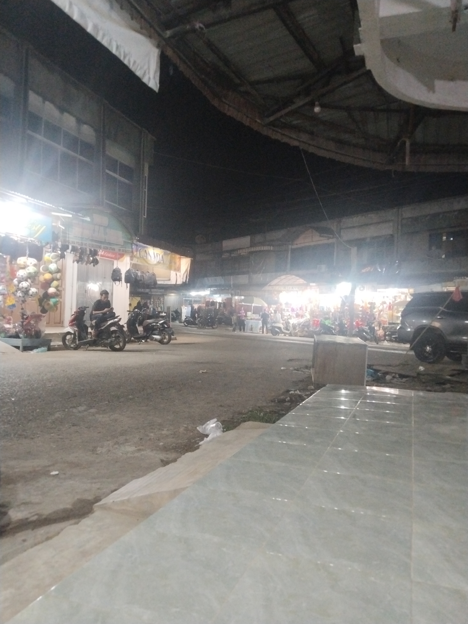 This nocturnal image captures a busy street scene in Keudegeudong, Indonesia, likely a market or commercial area. The sky is dark, indicating night, and the area is illuminated by various artificial lights from street lamps and numerous shopfronts.
In the foreground, a light-colored tiled walkway stretches diagonally across the bottom right, with a concrete curb separating it from the street. The street itself is asphalt or concrete, showing some wear.
On the left side of the street, several motorcycles are parked or ridden. One person is prominently seated on a white and red scooter, looking towards the right. Another person is visible further down the street, also on a motorcycle. Beyond them, a row of two-story buildings lines the street, their upper windows dimly visible. Shopfronts on the ground floor are brightly lit, displaying various merchandise such as hanging bags, what appear to be soccer balls, and other general goods.
In the mid-ground and extending to the right, more market stalls and shops are visible, bustling with activity. Several people can be seen moving around or standing near these stalls, suggesting a lively evening atmosphere. More motorcycles are parked in front of these shops. On the far right, a dark SUV is parked parallel to the street.
Overhead, a large, rustic metal roof structure, possibly part of a covered market or a wide awning, extends across the scene.
Visible text on shop signs includes "PELIMA POLISI NO. 12 KEUDEGEUDONG, ACEH UTARA" (an address or shop name), "HP. 0852627...", "CANADA" on a banner, and "APA Leather". The overall impression is that of a functional, active commercial hub after dark, likely catering to local residents.