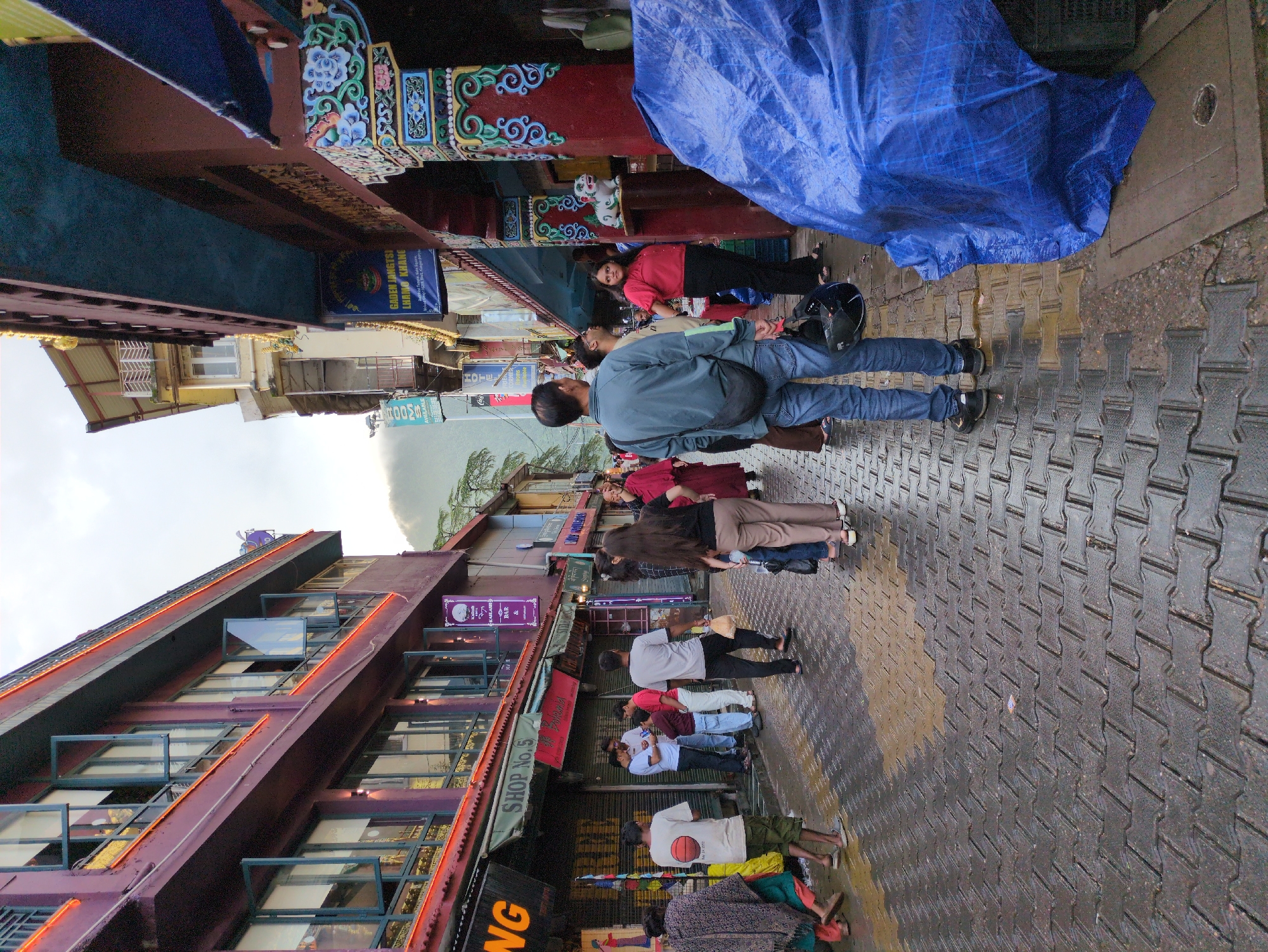 This image depicts a street scene in McLeod Ganj, Dharamshala, India, likely during or after rainfall. The street is paved with dark interlocking bricks, which appear wet and reflective. Buildings line both sides of the narrow street, with the left side featuring a prominent maroon-colored building with multiple windows and orange accent lighting. The right side shows a building with decorative colorful carvings on its columns and a blue awning.
Several people are present on the street. In the foreground, a man in a blue-grey jacket and jeans is walking away from the camera, holding a black helmet and a black bag. To his right, a large blue tarp covers an object or area. Further down the street, other individuals are seen walking or standing in small groups. Some are wearing traditional Tibetan attire, while others are in casual modern clothing. Signs are visible on the buildings, including one that reads "SHOP No. 5" and others with Tibetan script. The overall atmosphere suggests a commercial and bustling area, typical of a tourist destination. The sky is overcast, and a distant hillside is visible in the background.