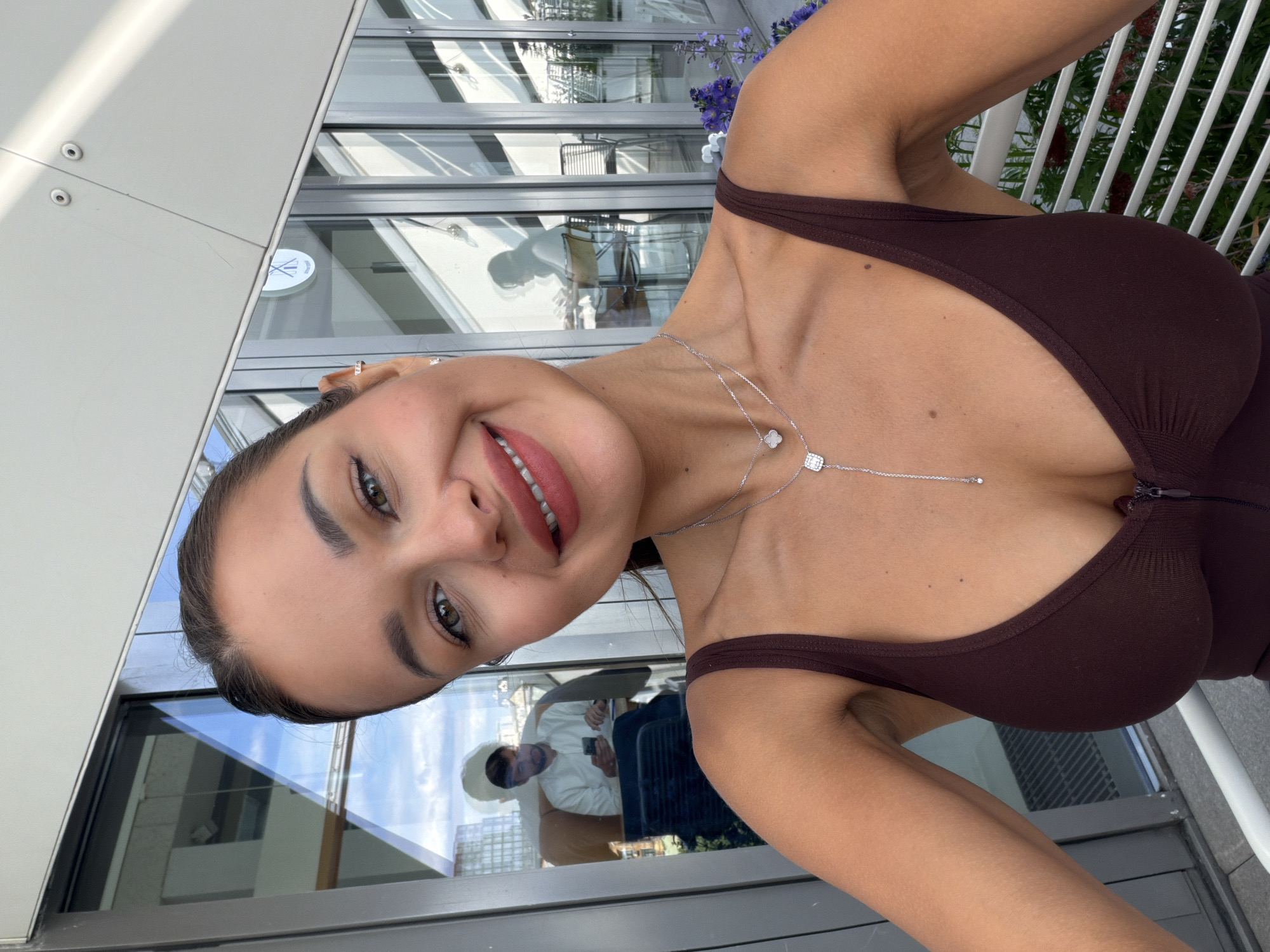 A smiling woman is captured in a close-up, angled selfie, seen from her upper chest upwards. She has dark hair pulled back, prominent eyebrows, and is looking towards the camera with a wide smile, revealing her teeth. Her lips are a muted pink. She is wearing a dark brown, possibly ribbed, tank top or bodysuit with thin straps. A delicate silver layered necklace adorns her neck and décolletage, featuring two small pendants: one a sparkling square and the other a four-leaf clover design. Small earrings are visible in her ears.
The background behind her features large, modern glass windows that reflect a bright, clear sky and contemporary building structures, suggesting an urban setting. Through the reflections, a man wearing a white long-sleeved top can be seen seated, appearing to look at a mobile phone. This suggests the woman is indoors, possibly at a café, restaurant, or lounge with a view. A white railing and some purple flowering plants are also visible through or reflected in the windows.
A circular white logo with the Roman numerals "XIX" is clearly reflected in one of the windows. The overall impression is of a bright, daytime scene. Given the context, this scene is set in Moscow, Russia.