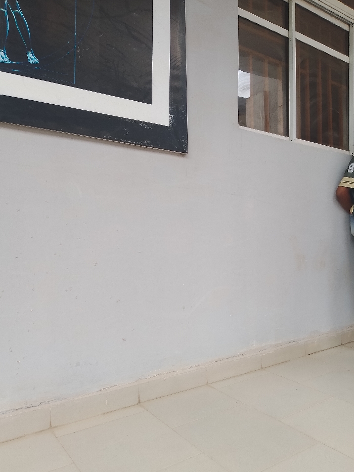 The scene appears to be an exterior shot, possibly along a building in Ogboko, Nigeria. The wall is painted a light gray color and has a large, framed picture on it. A window is visible to the right of the picture. In the lower part of the frame, there are light-colored tiles on the floor. A person is partially visible on the right side of the frame. The image focuses on the building's features, providing a simple documentation of the external facade.