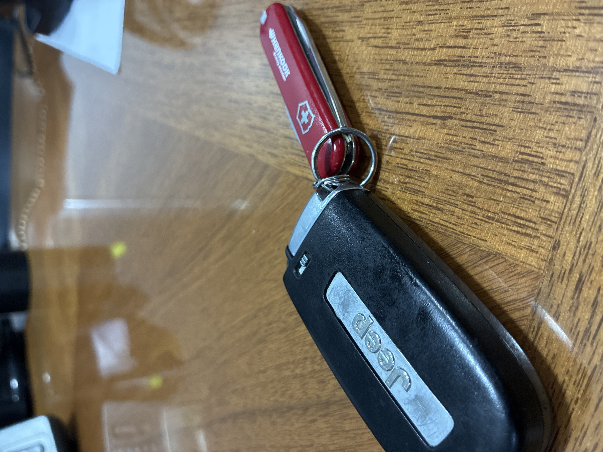 The image shows a Jeep car key and a red Hankook Swiss Army knife on a wooden surface. The key is black with a silver Jeep logo. The Swiss Army knife is attached to the key ring. The background is a brown wood surface. The location is unknown, and there is no evidence to suggest it is in Cuautitlán Izcalli, Mexico. There are no people visible.