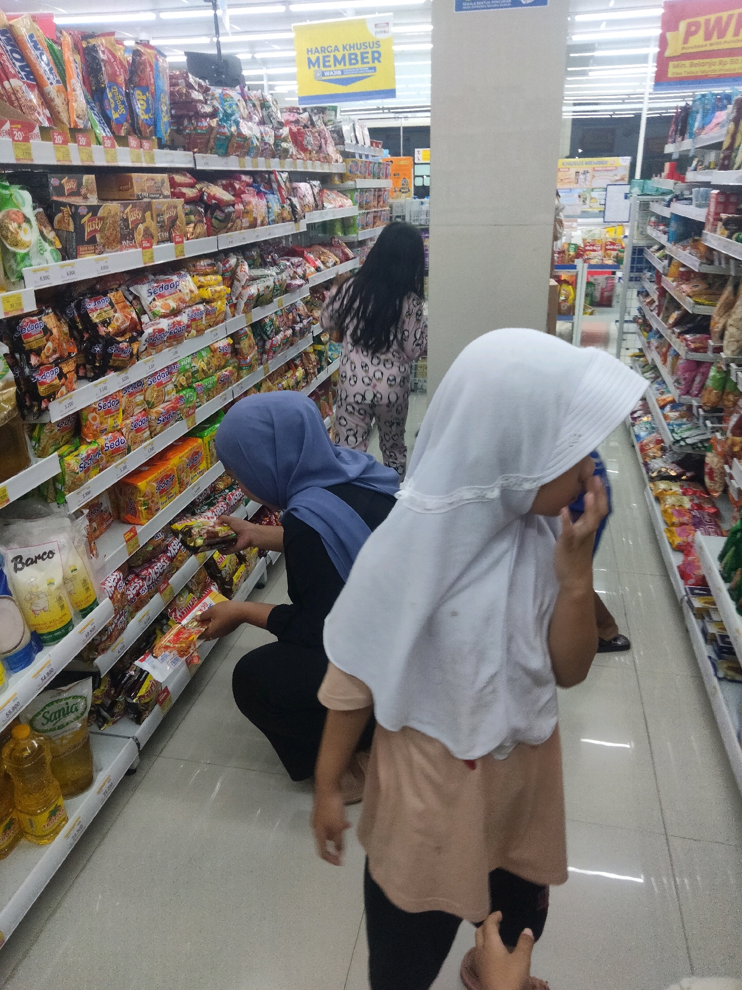 In the city of Suka Sari, Indonesia, a grocery store aisle is depicted. Two individuals, both female and wearing headscarves, are the primary subjects. One woman, wearing a blue headscarf and a black shirt, is crouched down, examining packages of instant noodles on a shelf. She holds two such packages in her hands. To her right, a younger female, wearing a white headscarf and a beige shirt over black pants, is partially visible, looking downwards and touching her face. In the background, another woman with long dark hair, wearing a patterned pajama-like outfit, walks away from the camera down the aisle. The shelves are densely stocked with various food products, including snacks, instant noodles, and bottles of cooking oil. The store appears well-lit with fluorescent ceiling lights. A promotional sign for "HARGA KHUSUS MEMBER" (Special Member Price) is visible above. The floor is tiled with a light-colored pattern.