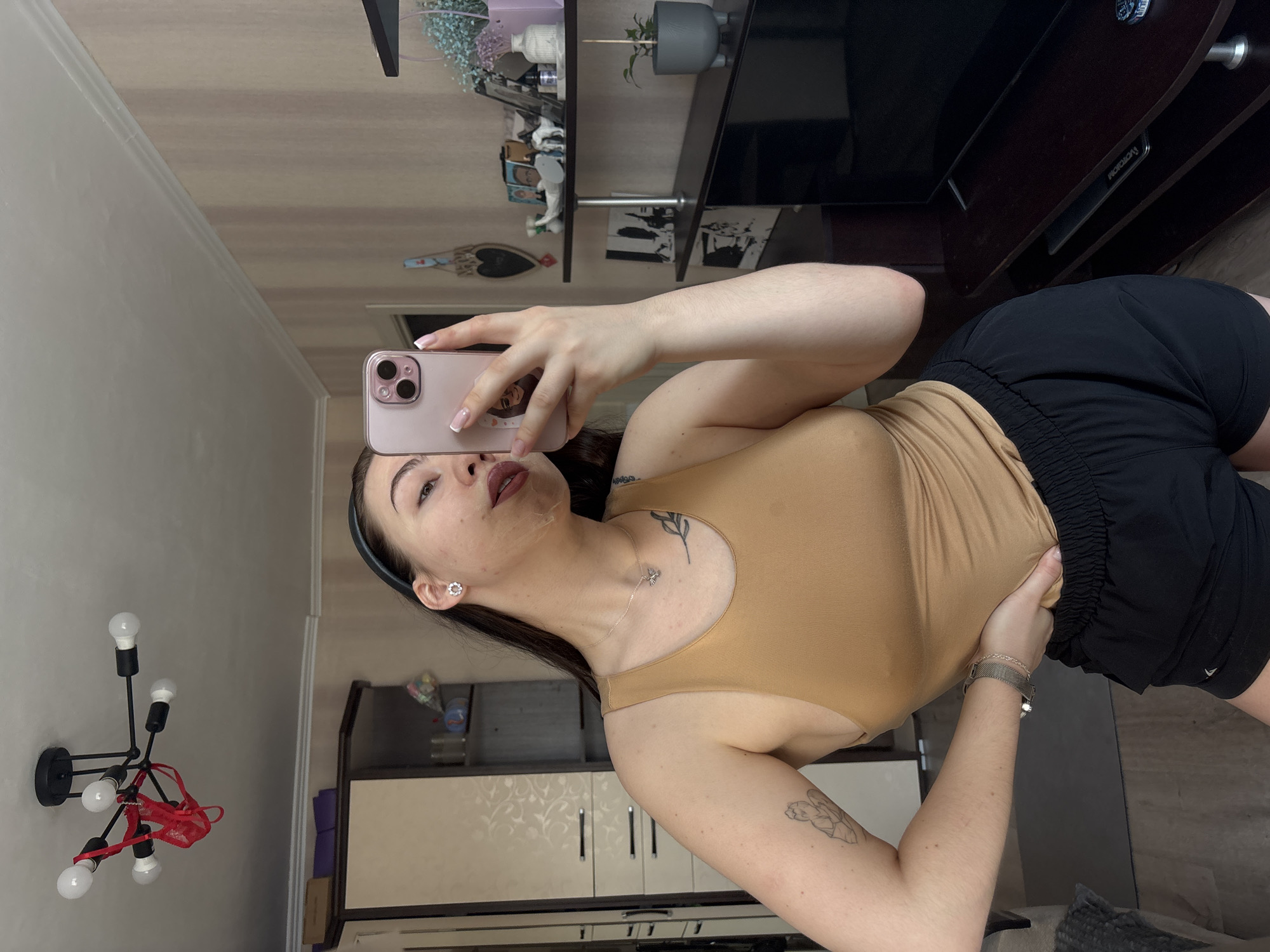 The image depicts a female subject taking a selfie within an indoor residential setting in Perm, Russia. The subject, an adult female with dark hair pulled back by a black headband, is positioned facing a reflective surface, holding a pink smartphone featuring a triple-lens camera array in her right hand. She wears a light brown tank top and black athletic shorts. Visible accessories include a silver-colored necklace with a butterfly pendant, small flower-shaped stud earrings, and a silver-colored mesh strap watch on her left wrist. Tattoos are present on her upper left arm (an outlined figure) and on her left clavicle area (a leafy design). Her left hand is placed on her left hip.
The background shows a room with light beige and pink striped wallpaper and white crown molding. A white ceiling features a black, multi-arm chandelier from which a red lace thong is suspended. To the left of the subject, a light-colored wooden wardrobe or cabinet with dark trim and horizontal handles is visible, with shelves holding various items. To the right, a dark brown entertainment unit displays a television (partially obscured) and shelves containing framed photos, small figurines (including a white horse), a potted plant, a vase with dried purple flowers, and a remote control. A heart-shaped decorative plaque hangs on the wall above the entertainment unit. The flooring appears to be light-colored wood or laminate, with a dark mat visible on the floor. The exact street-level location within Perm, Russia, cannot be determined from this indoor scene.