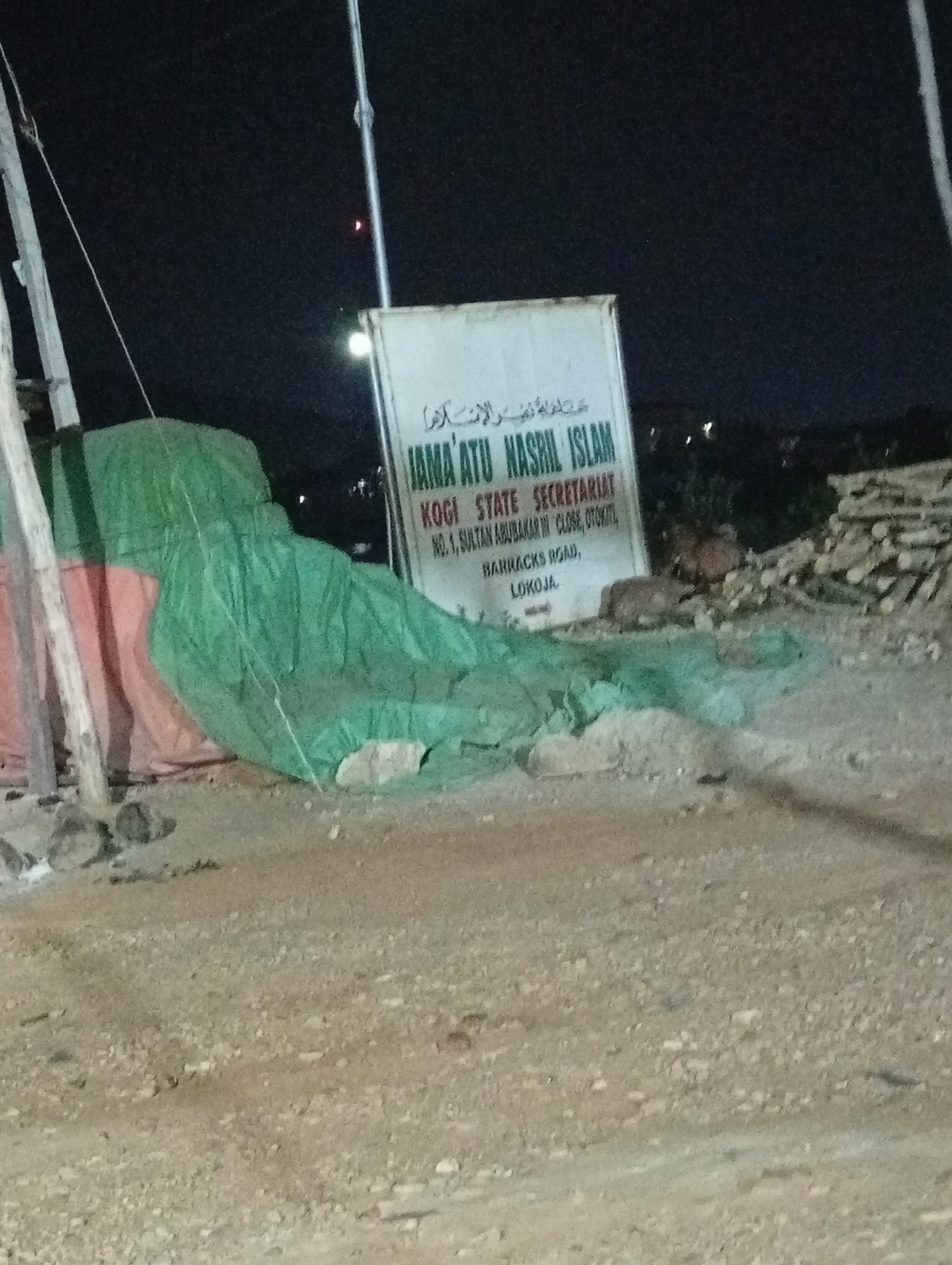 The image shows a sign for "Jama'atu Nasril Islam Kogi State Secretariat" located at No. 1, Sultan Abubakar III Close, Otoki, Barracks Road, Lokoja, Nigeria. The sign is positioned next to what appears to be construction materials covered by a green tarp. The photograph was taken at night.