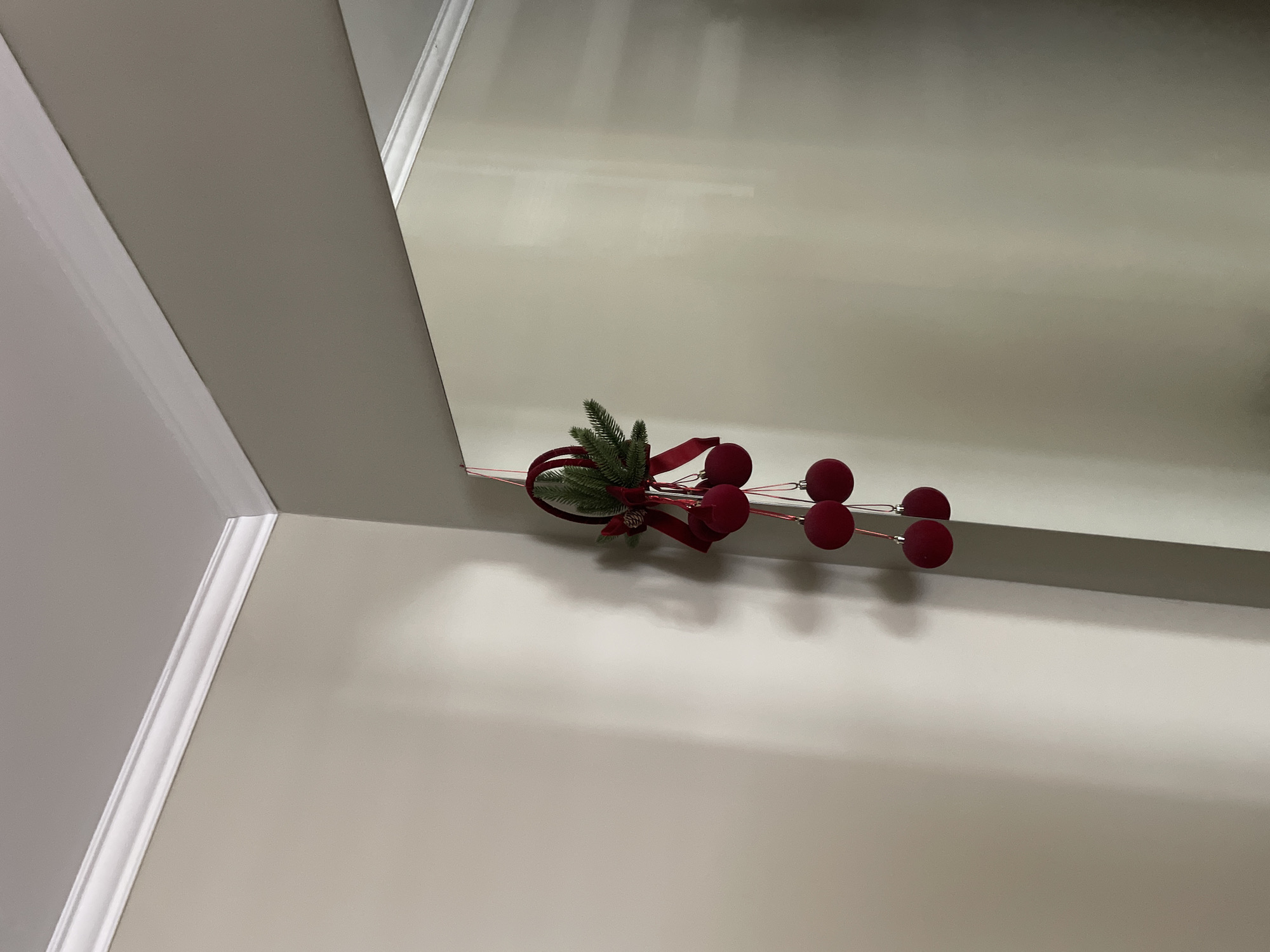 The image displays a Christmas decoration hanging from a corner where two walls meet. The decoration consists of greenery, red ribbons, and several red baubles. The walls are painted a neutral, light color and meet at a white crown molding. The lighting in the scene creates shadows on the walls, suggesting an indoor environment. There are no people visible in the image. The specific location in Cahul, Moldova, cannot be determined from the visual information provided.