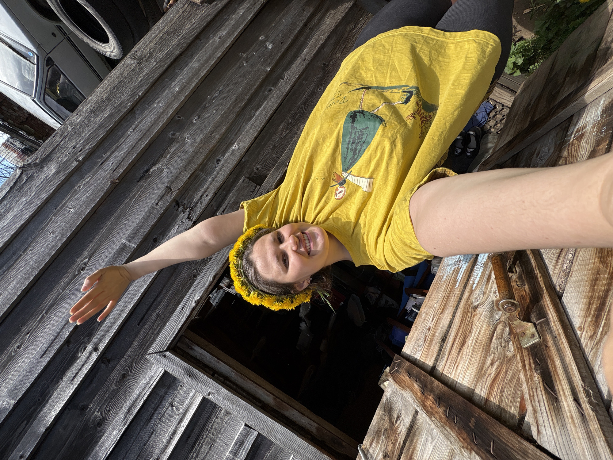 A young woman wearing a yellow t-shirt and a dandelion wreath on her head is seen in a selfie-style image. She is smiling and has one arm outstretched. The background consists of weathered wooden planks, suggesting a shed or similar structure. A car with tires visible is in the background. The location is unknown.