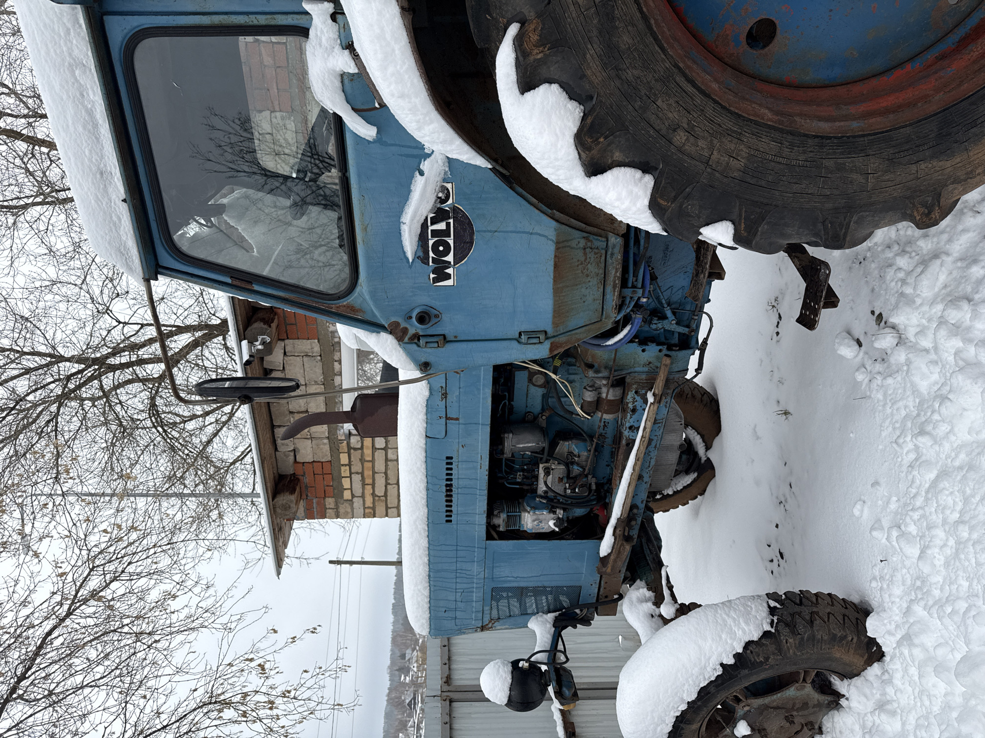 A blue Volvo tractor is partially covered in snow. The tractor is parked near a building made of brick and corrugated metal. The surrounding environment is snow-covered. Bare tree branches are visible in the background. The tire reads "MADE IN RUSSIA". It is located in the city of Luzhkovo, Russia. No people are present in the image.