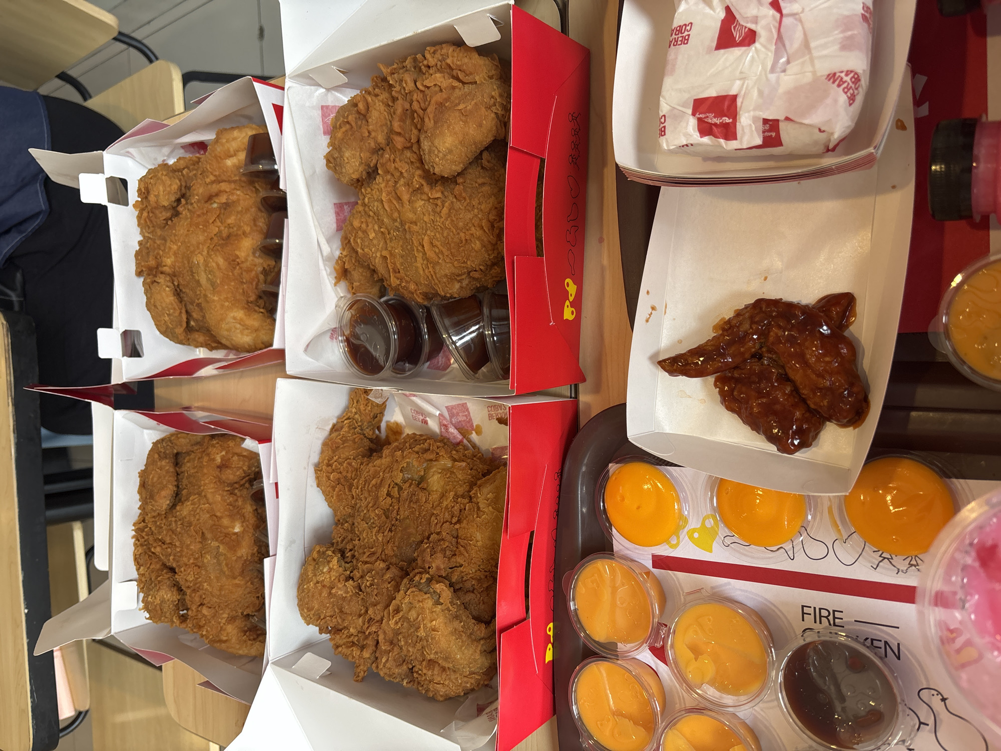 This image shows a table at a fast-food restaurant, likely a KFC outlet, located in Majalengka Kulon, Indonesia. The table is laden with several boxes of fried chicken, dipping sauces, and a packaged item, possibly a burger or wrap. In the foreground, there are small plastic containers filled with an orange sauce or puree, and some dark dipping sauces. The fried chicken is golden brown and appears to be a whole chicken or a significant portion of one in each box. One box in the lower right contains a white paper-wrapped item with red branding. The background shows wooden tables and chairs, suggesting a casual dining environment. The focus is on the food presentation, indicating a meal being served or about to be consumed.
