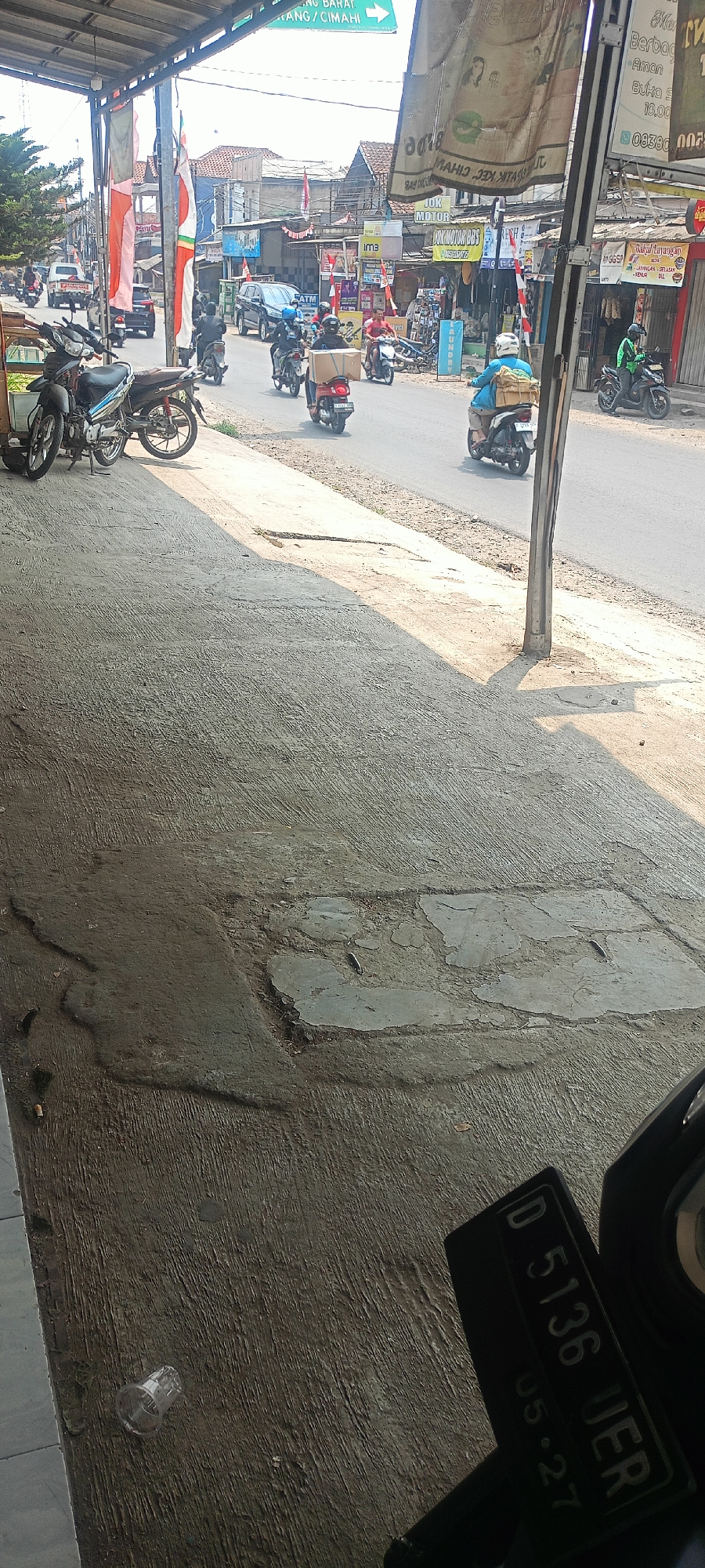 The image captures a street scene in Cipatik, Indonesia, on a sunny day. In the foreground, a grey concrete pavement is visible, with a few patches of darker, worn concrete and some debris, including cigarette butts and a small plastic cup. The pavement is partially shaded by an overhang structure on the left, where two parked motorcycles are present.
The main focus of the image is the street beyond the pavement. Several motorcycles are in motion on the asphalt road. On the left side of the road, a white car is seen, followed by other vehicles. A motorcycle with a rider wearing a blue shirt and carrying a large package strapped to the back is moving towards the right. Another motorcycle rider, wearing a red shirt and also carrying a box, is ahead of them. Further down the road, a motorcycle with a rider in a green jacket is visible.
The background consists of buildings lining the street, with various shop signs and flags displayed. Some of these signs are in Indonesian. The sky is bright and clear, indicating a daytime scene. The overall impression is of a busy street in a commercial area of Cipatik.