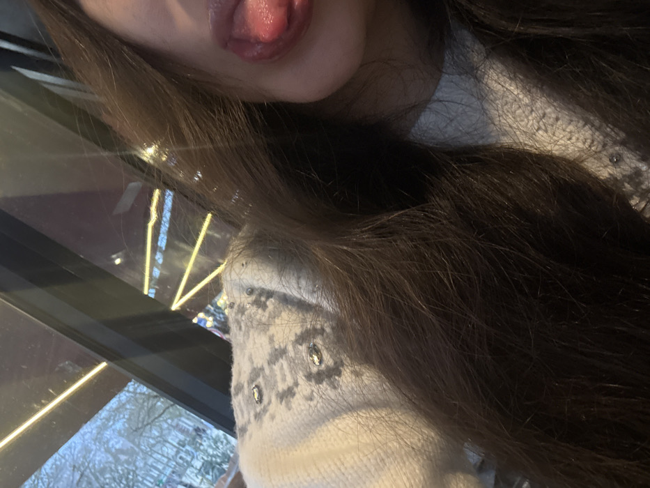A person sticks their tongue out in a selfie. Long, dark brown hair dominates the image. The individual wears a white sweater or garment with a fair isle pattern in gray, accentuated by decorative studs or beads. Reflections of light and a cityscape can be seen through a window beside the person.